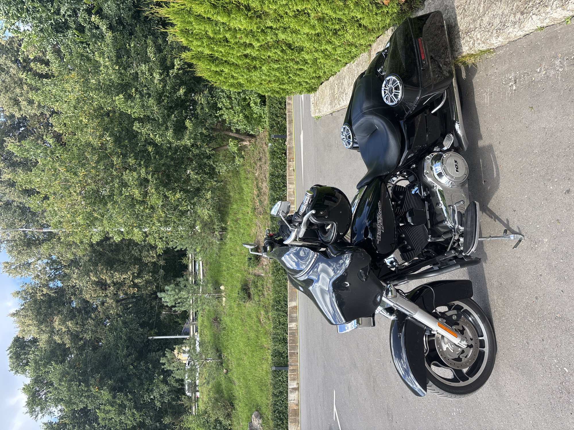 A black Harley-Davidson motorcycle is parked on an asphalt road adjacent to a manicured green hedge. The motorcycle is positioned facing left, with its front wheel turned slightly inward. The bike features chrome accents, a large fairing, and saddlebags. Two speakers are visible on the rear of the motorcycle. Behind the hedge, a grassy incline is visible, dotted with several small evergreen trees. Further in the background, a dense line of mature trees forms a natural backdrop, with a bright blue sky visible above. The scene appears to be an outdoor setting, possibly a roadside or a park area, given the presence of both the paved road and the natural landscaping. The specific location is the city of Zvenigorod, Russia. There are no people present in the image.