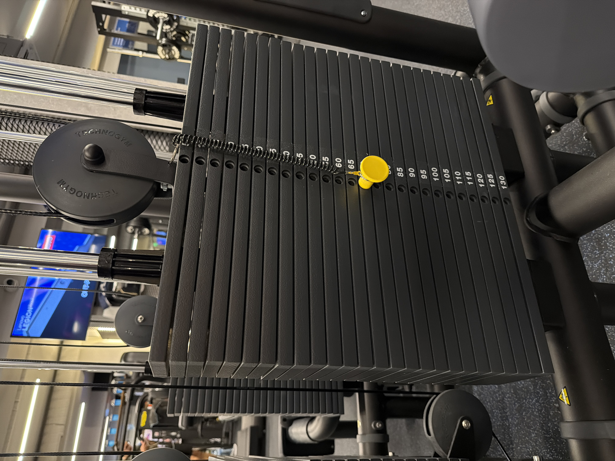 The image shows a weight stack on a Technogym exercise machine. The weights are black metal plates stacked vertically. The weight indicator pin, which appears to be yellow plastic, is inserted at the 70 weight mark. The numbers indicating the weight values are printed in white on the plates. The brand name Technogym is visible on a pulley wheel on the left side of the stack. The location appears to be a commercial gym with other equipment visible in the background, however, the precise location in Varna, Bulgaria cannot be determined from the image. There are no people visible in the frame.