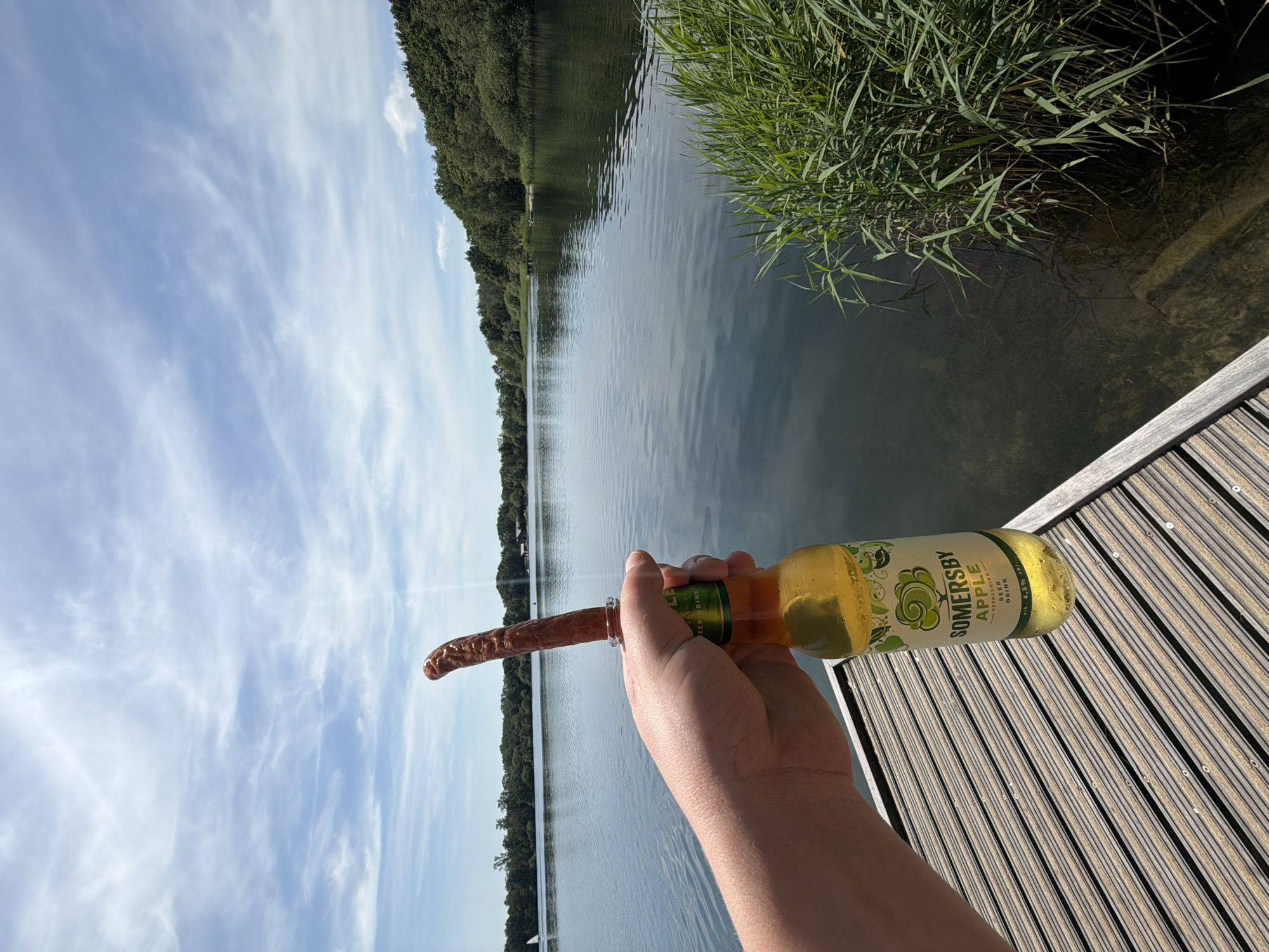 A first-person perspective shot captures a hand holding a bottle of Somersby Apple beer, with a long, thin sausage inserted into the bottle's opening, positioned near a body of water. The background reveals a serene lake surrounded by lush green trees under a partly cloudy sky. A wooden deck is visible in the foreground, suggesting a lakeside setting, possibly in the city of Turnhout, Belgium, given the context of the surrounding scenery. The water is calm, with ripples suggesting a light breeze. The overall impression is one of a leisurely outdoor activity.