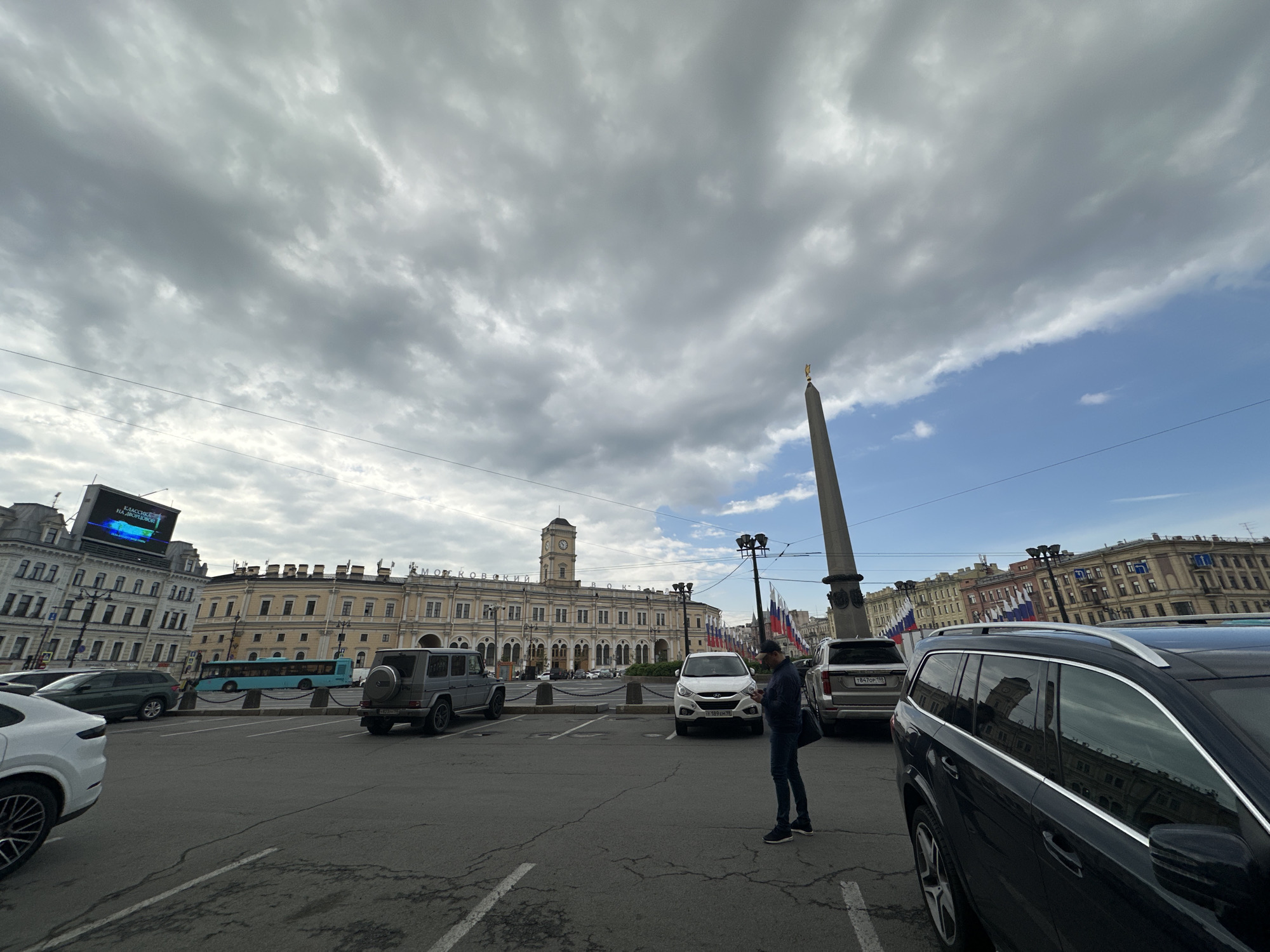The image depicts a scene in Saint Petersburg, Russia, specifically at Ploshchad Vosstaniya (Uprising Square) in front of the Moskovsky railway station. The Alexandrian Column can be seen in the background. The foreground shows a parking lot with several cars. A man dressed in jeans and a dark top is standing in the lot, facing away from the camera. The sky is overcast with heavy clouds, though patches of blue are visible. The overall environment appears urban with architecture typical of Saint Petersburg.
