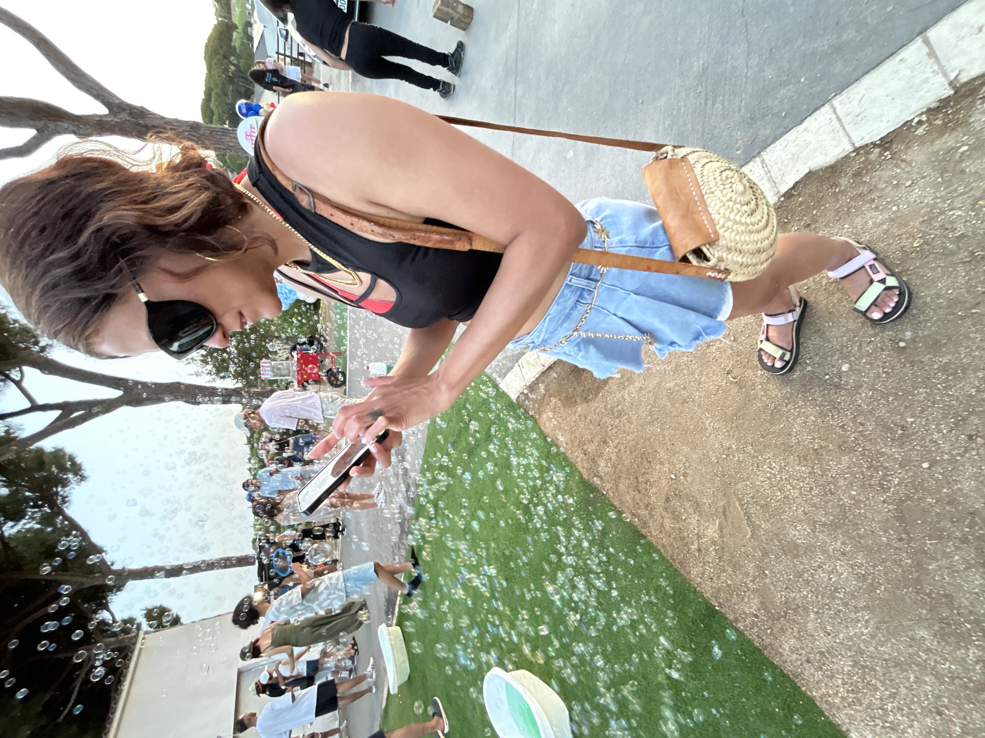 An outdoor scene with many bubbles floating in the air. In the foreground, a woman is looking at her smartphone. She is wearing a black crop top, a denim skirt, and colorful sandals. She has a straw bag slung over her shoulder. In the background, other people are visible, some walking and some interacting with a bubble machine. There are trees and a building in the background, and the ground is a mix of gravel and artificial turf. This event is taking place in Sant Vicenç De Montalt, Spain.