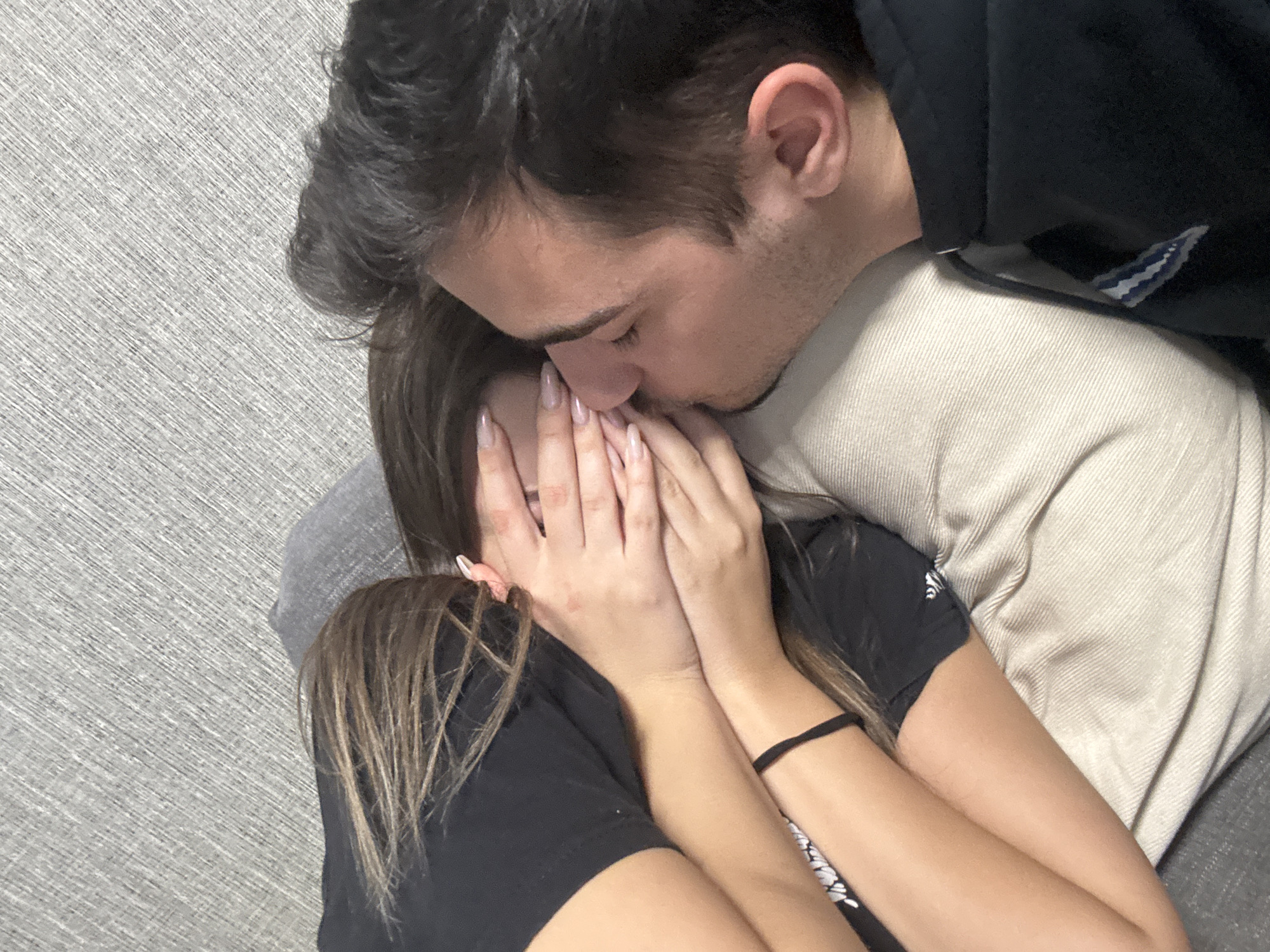 A man kisses a woman, who is covering her face with her hands. The man is wearing a beige shirt and a black jacket, and the woman is wearing a black t-shirt. They are indoors, leaning back against a gray textured wall. The location in Sofia, Bulgaria is not possible to determine from the provided image.
