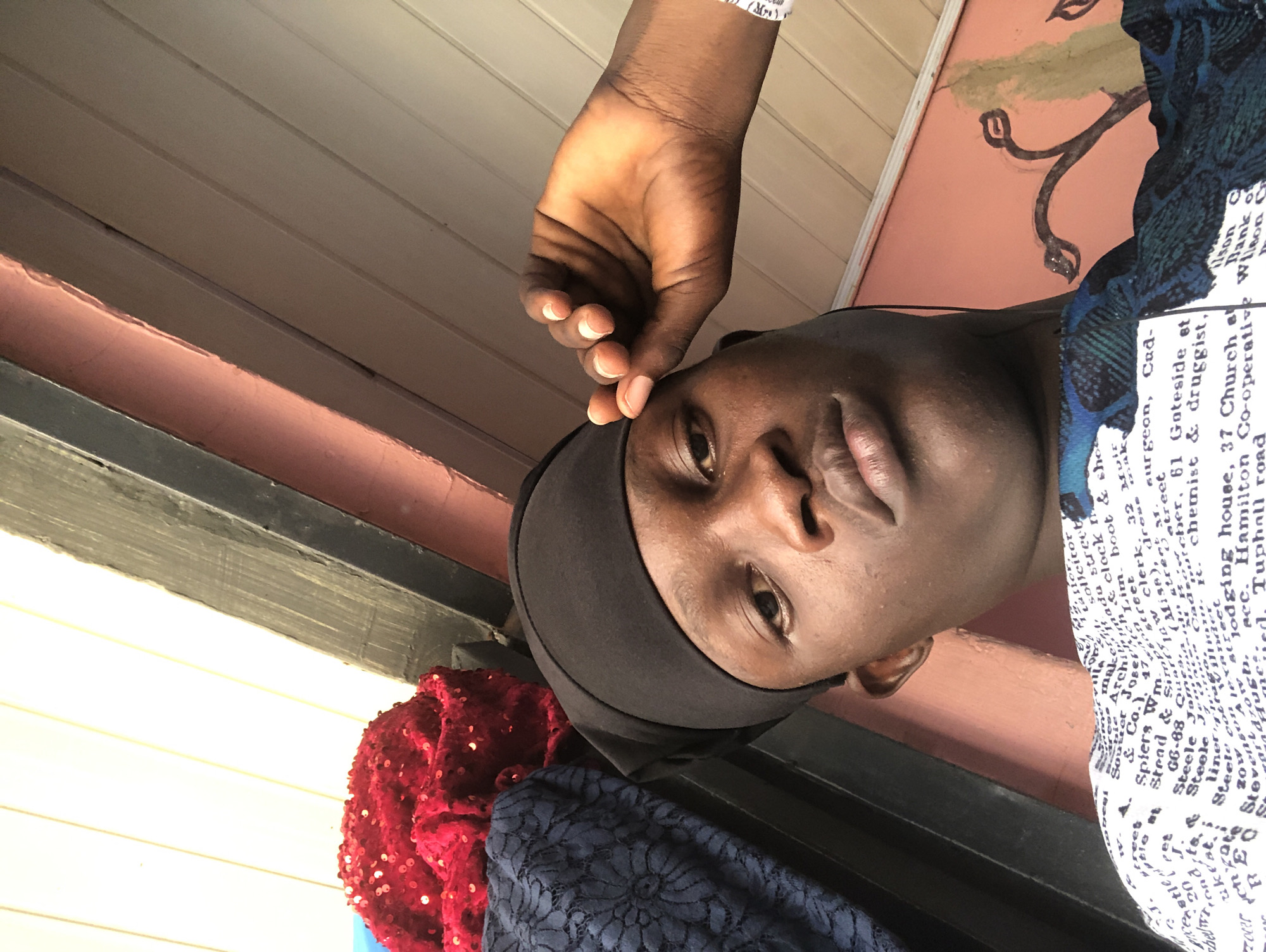 A close-up, angled selfie captures a young Black male with dark skin and short hair, wearing a black head covering and a patterned blue and green shirt. He is looking directly at the camera with a neutral expression. His right hand is raised, touching his forehead. In the background, a red sequined object and a dark blue lace fabric are visible. The wall behind him is painted a light pink hue and has what appears to be a faded mural of a plant. The ceiling is made of parallel wooden planks. The image composition and visible elements suggest a casual, indoor setting possibly in Ado, Nigeria, given the provided context of the location in the prompt.