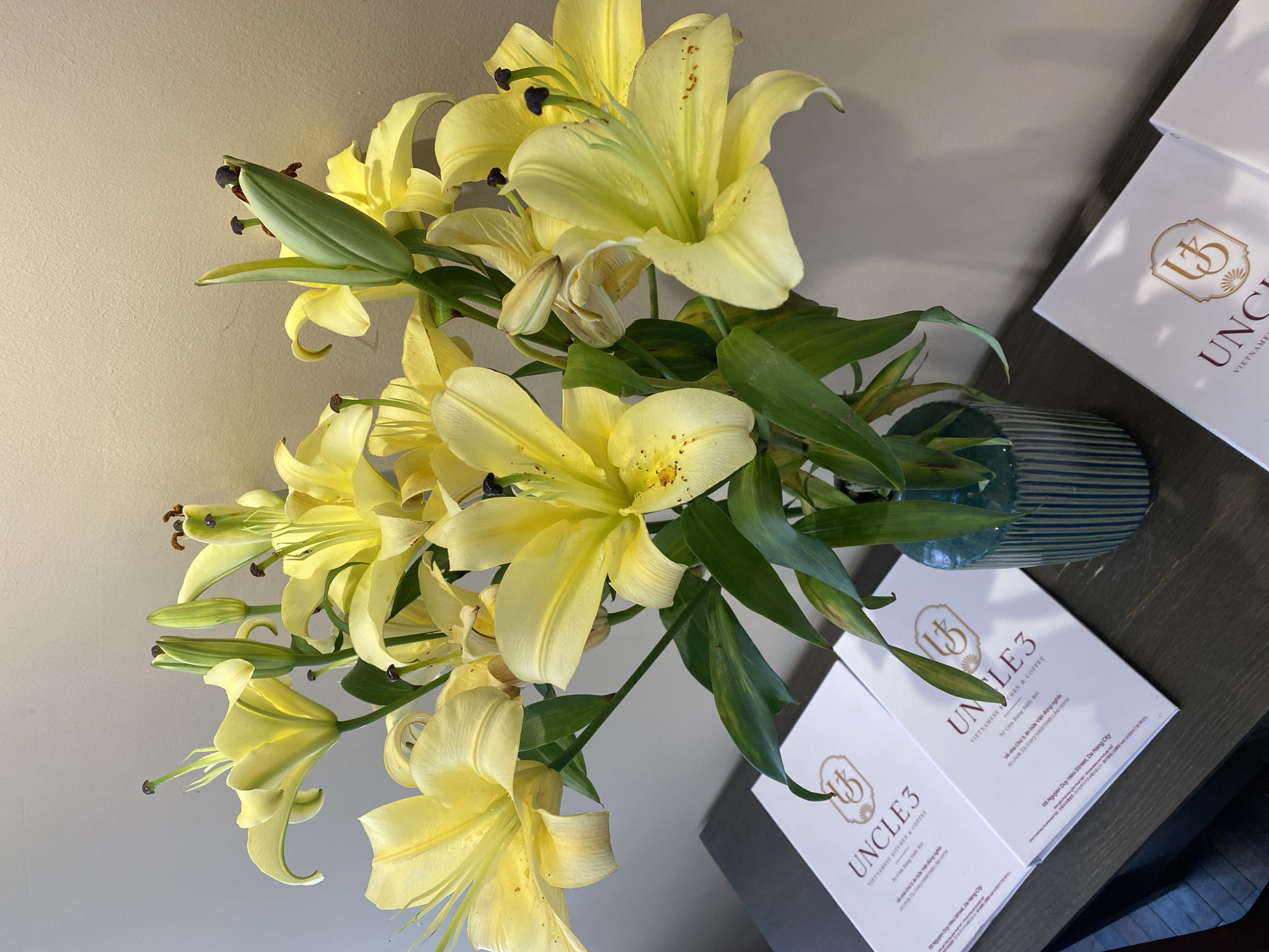 A vase of yellow lilies is displayed on a dark wooden surface next to three white brochures. The brochures feature a golden logo and text that reads "UNCLE 3 VIETNAMESE KITCHEN & COFFEE" and an address in Da Nang, Vietnam. The lilies are in various stages of bloom, with some fully open and others still in bud. The background is a neutral-toned wall.