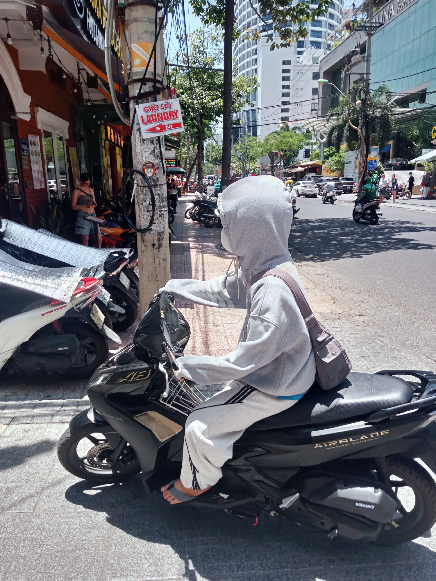 The image captures a street scene in Nha Trang, Vietnam. In the foreground, a person wearing a gray hoodie, white sweatpants, and a face mask is seated on a black Honda Air Blade scooter. The person has a crossbody bag. The scooter is parked on the sidewalk next to other motorcycles covered with a silver protective cover. Behind the scooter, a utility pole with a "Laundry 5M" sign is visible. In the background, the busy street is filled with cars, motorcycles, and pedestrians. Buildings, including the "Galina Hotel", are visible in the background, indicating an urban environment. A laundry establishment is nearby. A woman with short shorts is looking at her phone in front of a store.