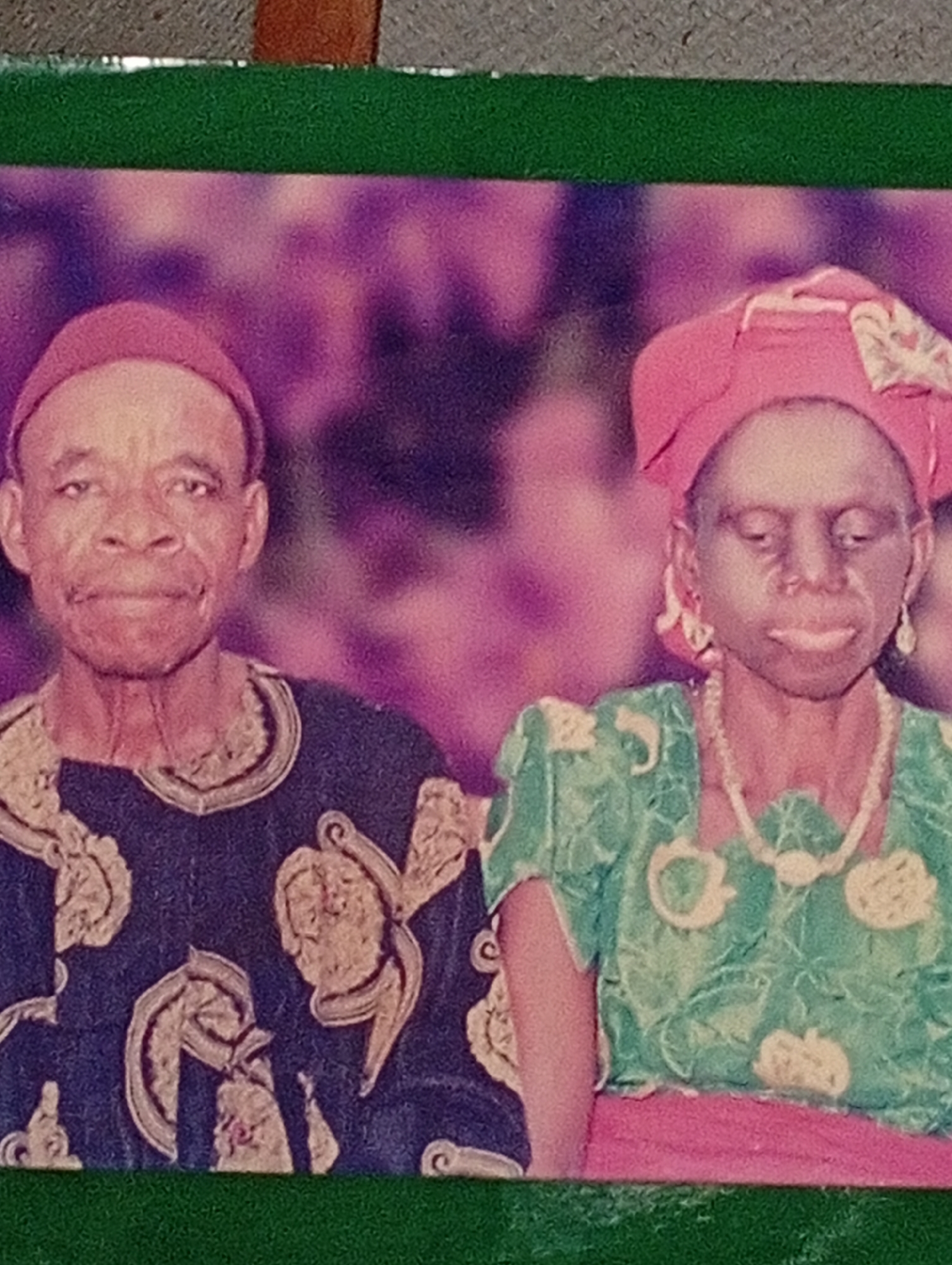 An undated, medium-shot, color photograph shows a portrait of an elderly African couple, a man and a woman, posed outdoors. The couple faces forward, with the man on the left and the woman on the right. Both are wearing traditional attire. The man wears a dark blue, long-sleeved garment with intricate gold embroidery around the neckline and cuffs, and a red cap. The woman wears a green dress with floral patterns and a pink headwrap. She also has on dangling earrings and a pearl necklace. The background is a blur of purple and pink, suggesting a festive or celebratory event. The photograph appears to be a studio portrait, or taken during a formal occasion. The location is Adda, Nigeria.
