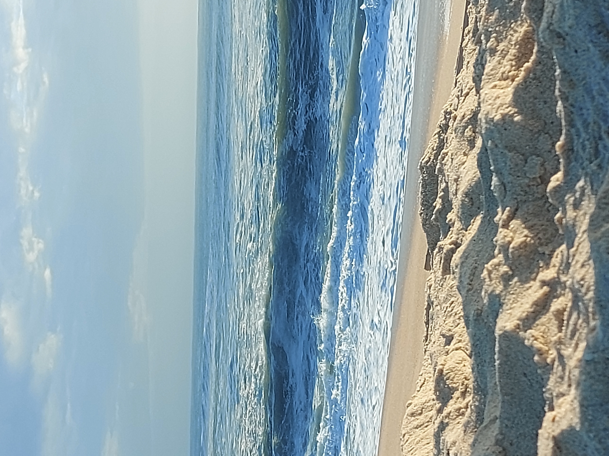 The image captures a scene at a beach in Figueira da Foz, Portugal. It shows the shoreline with sand on the right, the ocean water in the middle, and the sky with some clouds on the left. The waves are visible in the water, and the sand appears to be textured with various formations. The lighting suggests it is daytime.