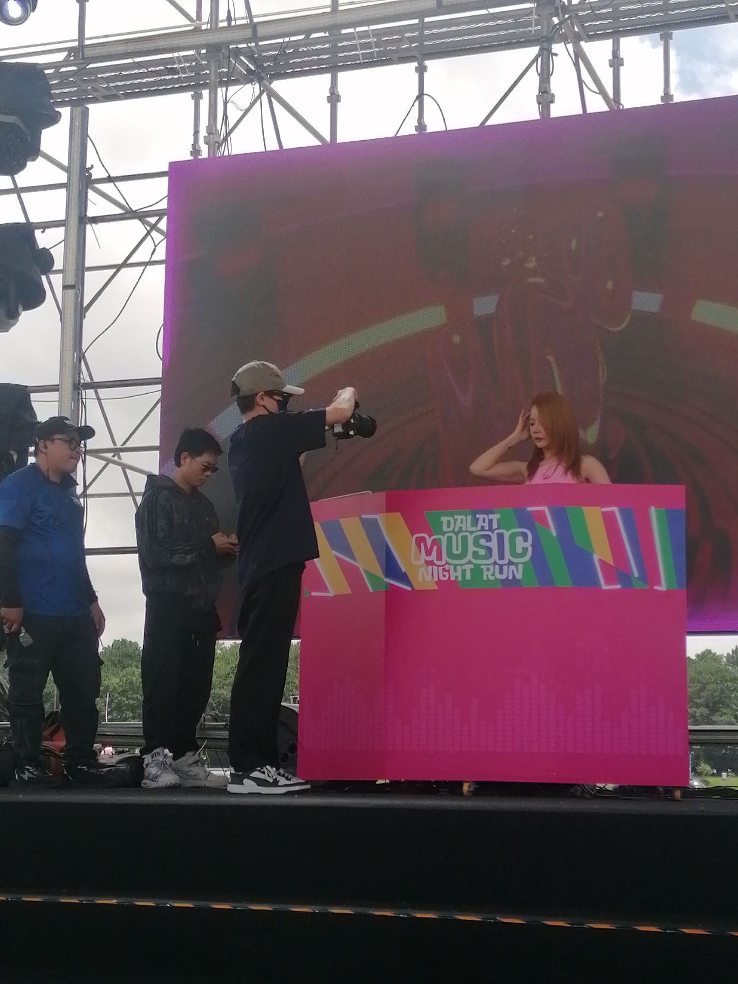 The image captures an outdoor stage setup during the "Dalat Music Night Run" event in Da Lat, Vietnam. Four individuals are present on the elevated black stage. On the right, a female with long brown hair, wearing a pink sleeveless top, stands behind a bright pink DJ booth. Her left hand is raised towards her head. The front of the DJ booth displays a multi-colored banner with the text "DALAT MUSIC NIGHT RUN" and abstract geometric patterns, along with a faint graphic equalizer pattern on its lower half. Behind the DJ booth, a large rectangular LED screen displays abstract red and purple graphic designs. To the left of the DJ booth, a man wearing a gray baseball cap, sunglasses, and a dark face mask holds a black professional camera, aiming it towards the female performer. Further left, another man in a black hooded jacket and sunglasses is looking down at a smartphone in his hands. To his left, a third man wearing a blue short-sleeved polo shirt over a dark long-sleeved shirt and glasses holds a small, dark rectangular object in his left hand while observing the scene. Above the stage, metal trussing structures supporting black stage lights are visible against an overcast sky. Green trees are discernible in the distant background behind the stage.