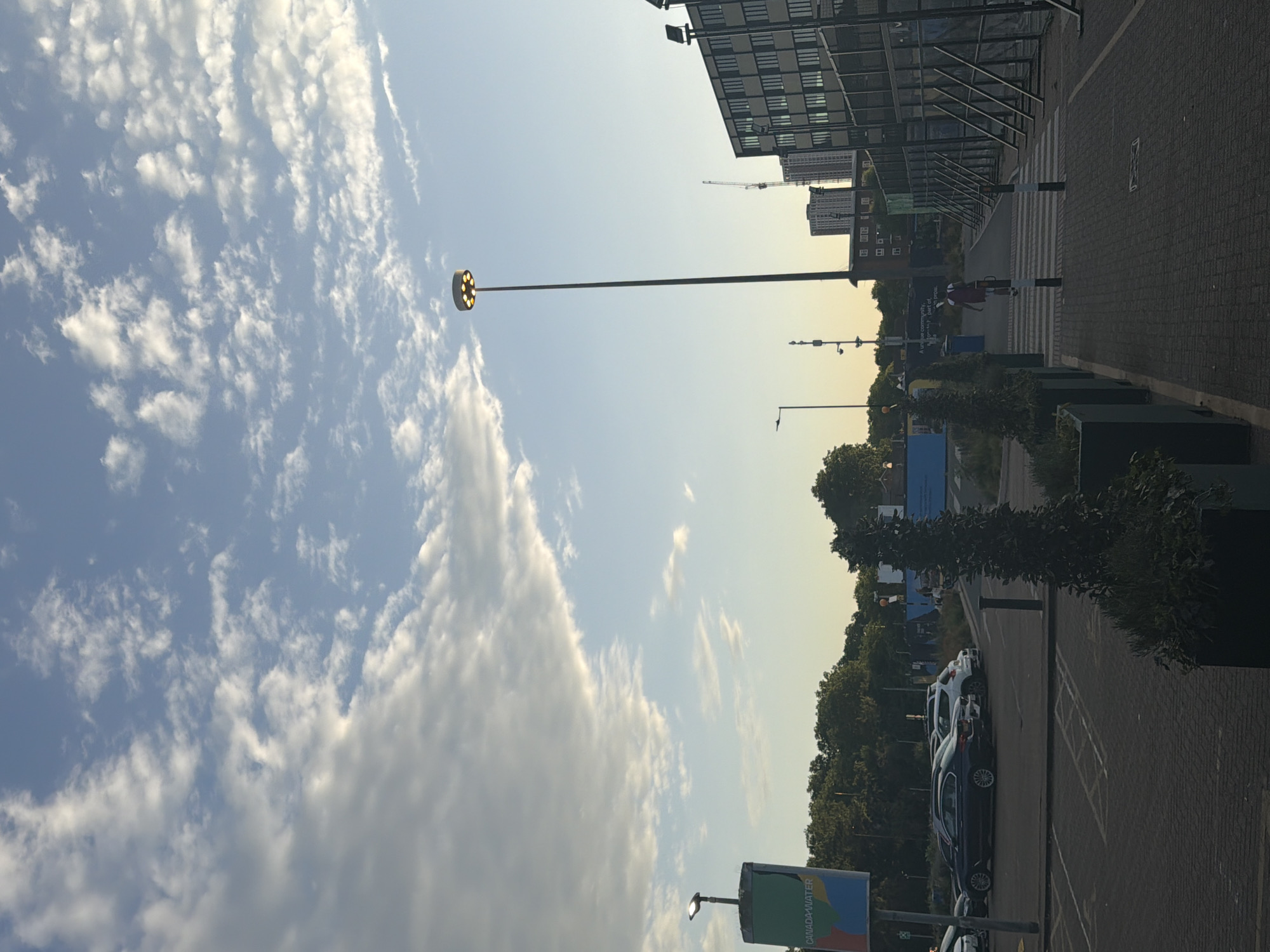 The image captures a scene in London, United Kingdom, possibly in a commercial or residential area. The sky is partly cloudy, with a tall lamppost featuring a ring of lights. The perspective is looking up, with buildings lining the right side of the frame. A walkway with zebra crossing is visible. A line of cars parked alongside a row of potted plants. A sign with “CANADA-WATER” is in the foreground. A person in red clothing is crossing the walkway. A construction crane is visible in the background, near other buildings.