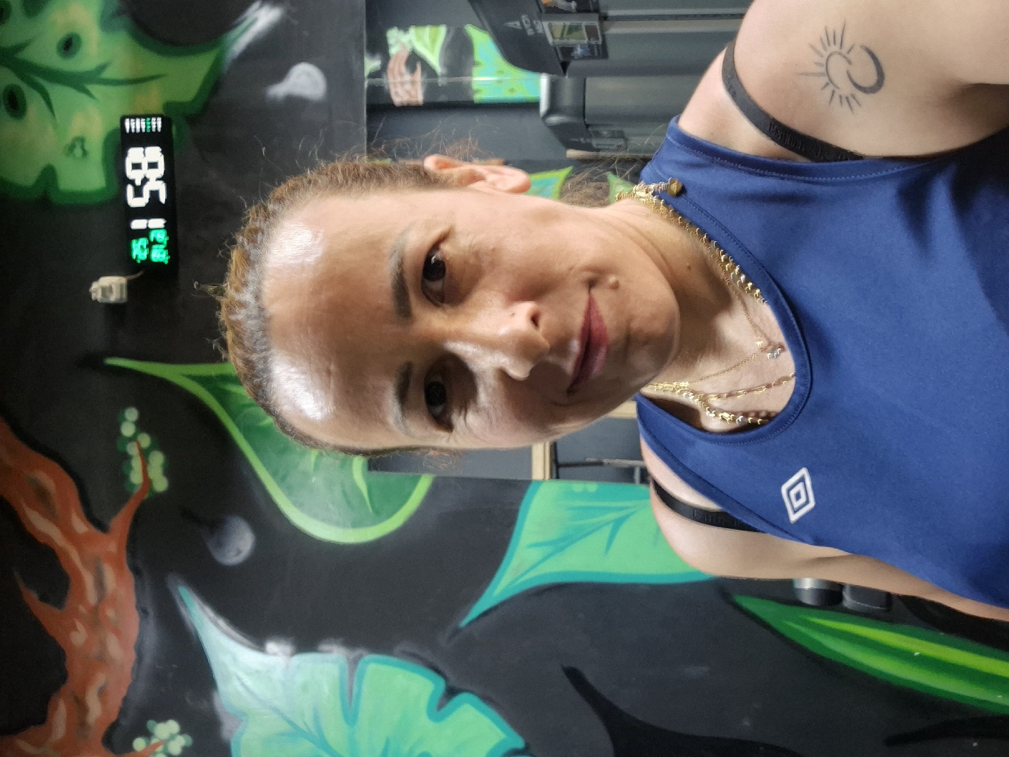 The image shows a woman posing for a selfie. She is wearing a blue tank top with a white diamond logo on it and several gold necklaces. She has a tattoo of a sun and moon on her upper right arm. The background appears to be a wall with graffiti art depicting green leaves and an orange tree. To the left of the woman, a digital display shows "85 | 1". The lighting suggests an indoor setting, possibly a gym or a studio. The woman's expression is a slight smile, and she seems confident and relaxed. The overall mood is casual and personal.