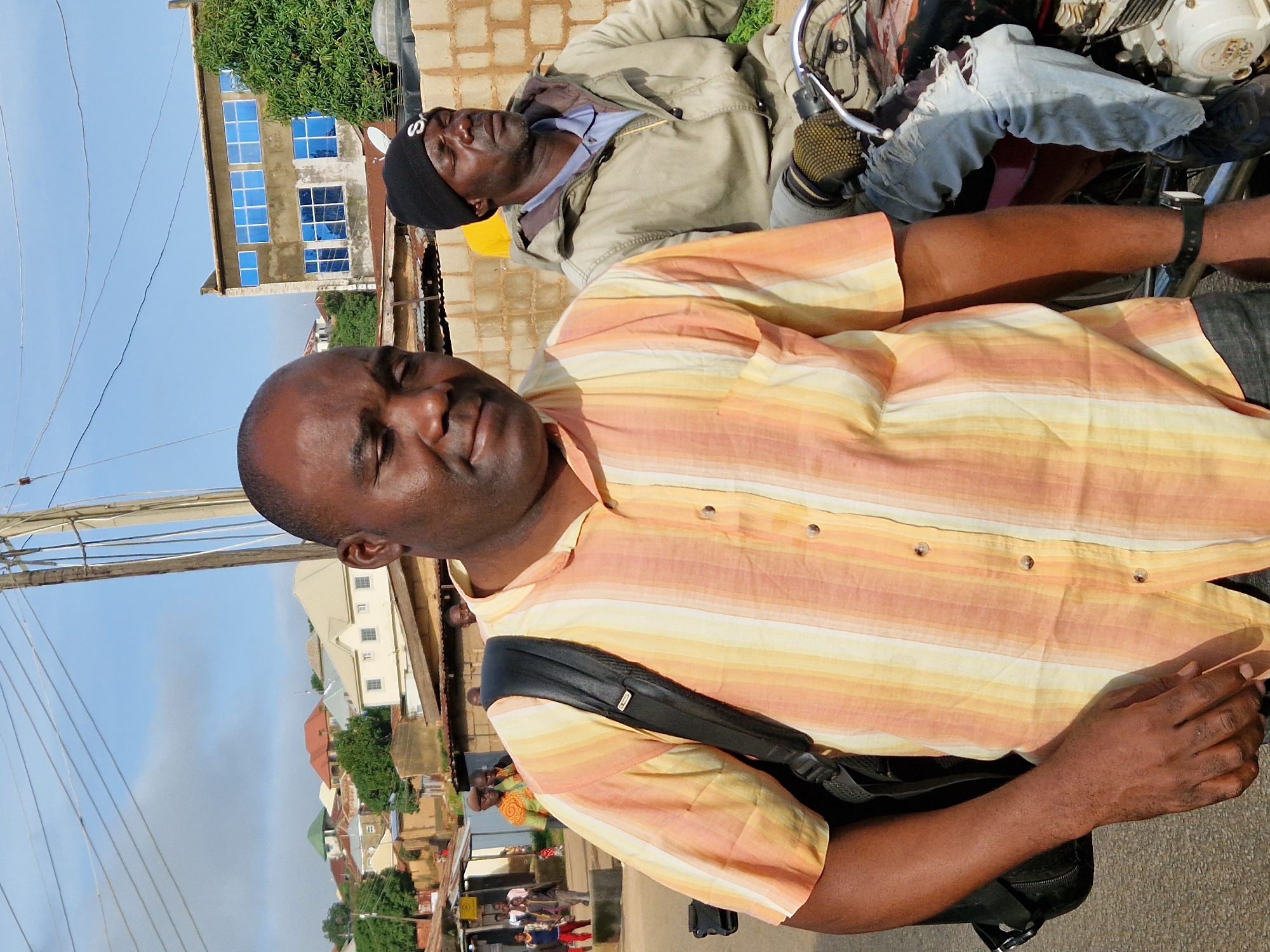 In Kabong, Nigeria, two men are pictured on a street with a building in the background. The man in the foreground, with dark skin and wearing a striped orange and yellow button-down shirt, carries a black backpack over his left shoulder. His gaze is directed downwards, and his right hand is visible, resting on his hip. To his right, a man with dark skin, wearing a black beanie and a tan jacket over a blue shirt, sits on a motorcycle. His eyes are closed. The motorcycle is partially visible, with its engine and handlebars in focus. The background shows a brick wall, a tree, and a multi-story building with blue windows. A power line stretches across the sky. Other people and structures are visible further in the background, suggesting a residential or commercial area.