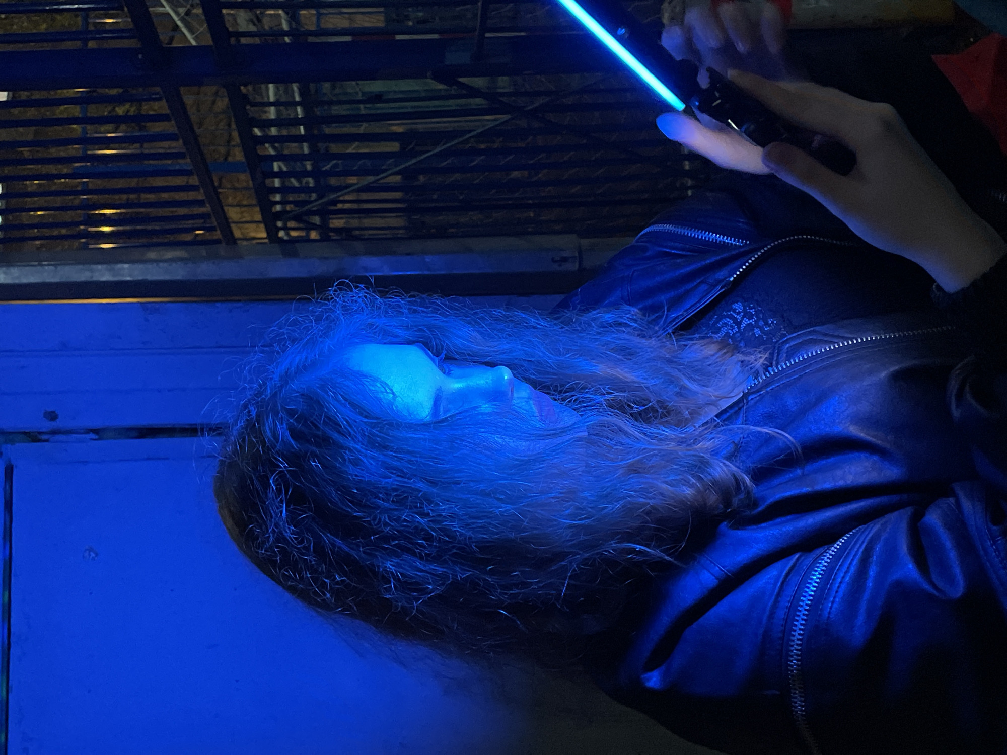 A side-view, medium shot shows a person with light hair illuminated in blue light, looking at a handheld device. The person is wearing a dark leather jacket. In the background, there is a fire escape and a wall. The scene is likely set outdoors at night. There is no information to suggest the photo was taken in Moscow, Russia.