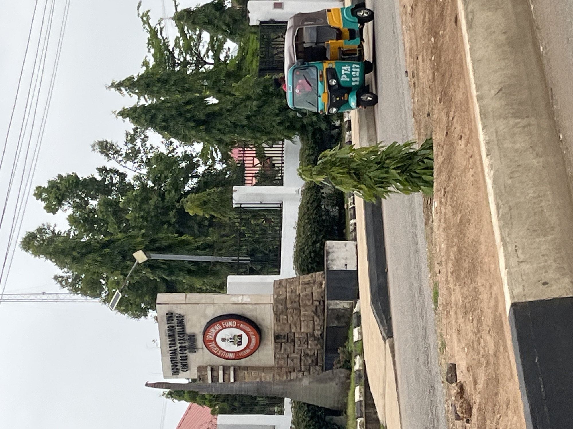 The image shows the entrance to the Industrial Training Fund (ITF) Centre for Excellence in Bukuru, Nigeria. A signpost with "INDUSTRIAL TRAINING FUND CENTRE FOR EXCELLENCE BUKURU" is prominently displayed, along with the ITF logo. To the right of the signpost, a teal and yellow tricycle, also known as an 'okada' or 'keke napep', with the license plate "PTA 11217" is parked on the side of the road. The tricycle appears to be occupied by at least one person, though their features are not clearly discernible. In the background, lush green trees and a fence line the property. The road in the foreground is paved, with a clear delineation between the road surface and the sandy embankment. Overhead, power lines are visible against a cloudy sky. The overall scene depicts a public institution and its immediate surroundings in an urban environment.