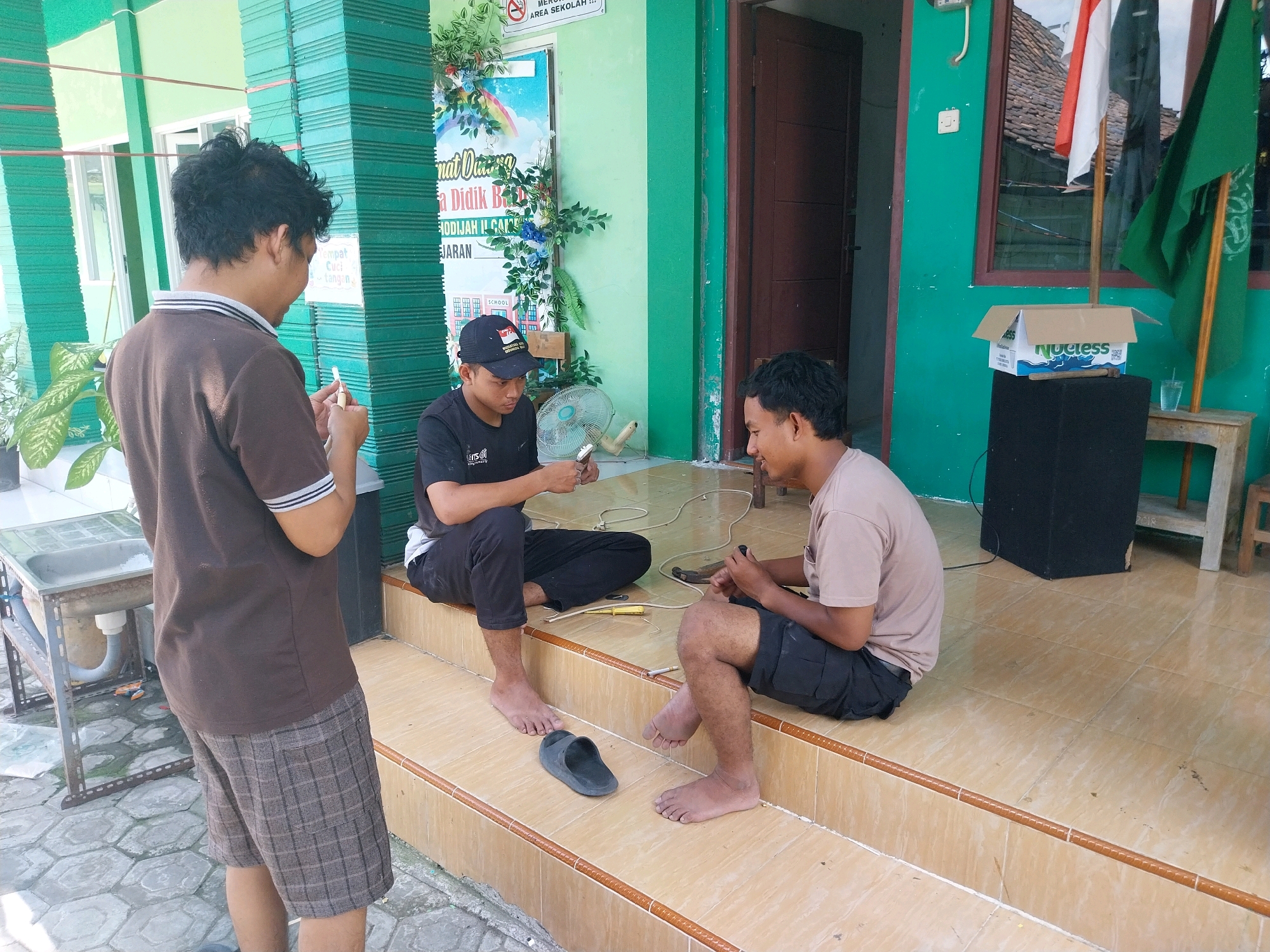 In Campur, Indonesia, three young men are seated outside a building. On the left, a man wearing a brown t-shirt and plaid shorts stands, holding a cigarette. In the center, a man in a black t-shirt and black pants sits on the stairs, looking at his phone. To his right, another man in a tan t-shirt and black shorts also sits on the stairs, holding a phone. A small oscillating fan is placed on the ground near the man in the black t-shirt. Behind them, a green building with white windows and a brown door is visible. Several flags are mounted on poles in front of the building. A cardboard box with "Nuefess" branding sits on a black speaker to the right of the men. A glass of water is on a small wooden table next to the speaker. The ground is tiled with hexagonal pavers, and the stairs are made of light-colored tiles with brown trim.
