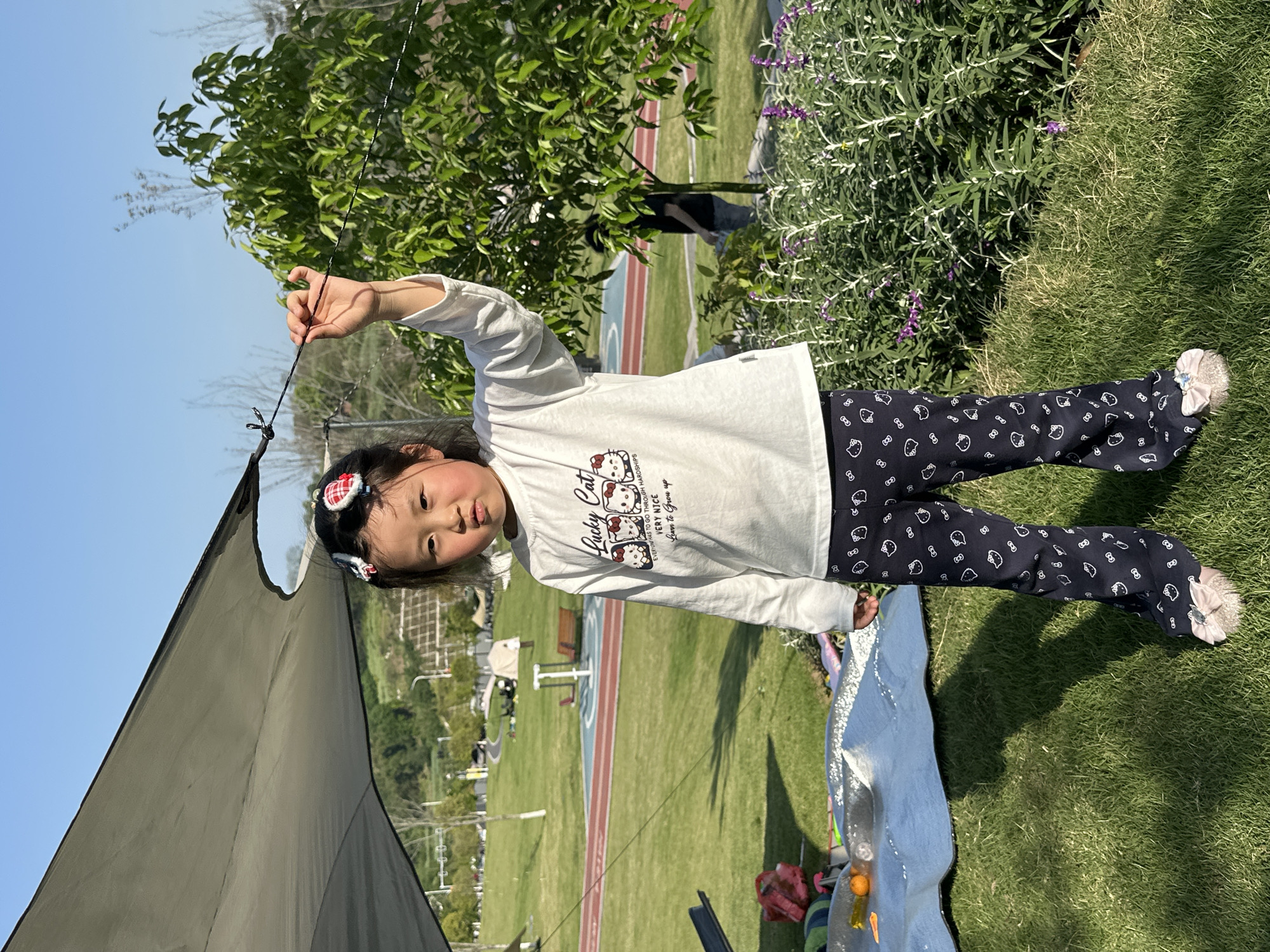 A young girl stands on the grass in a park. She is wearing a white long-sleeved shirt with a Hello Kitty design and patterned leggings. She is holding a black tent cord in her right hand and looking at the camera. Behind her, there is a green bush with purple flowers, a tree, and a park scene with a running track and a sports field. A green tent corner is visible on the left. There is a blanket, bottles, and fruit on the grass in the lower center of the image. The photograph appears to have been taken in Jiangjin, China.