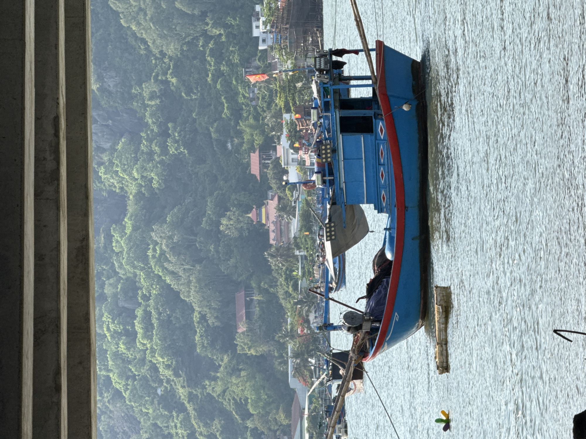 This is a medium shot of a blue and red fishing boat anchored in the water, with a lush green hillside covered in trees and vegetation in the background. The boat is in the foreground, taking up a significant portion of the frame. It has a cabin structure and various fishing equipment. A small, colorful buoy and a piece of what appears to be styrofoam float in the water near the boat. In the middle ground, there is a small settlement with buildings, some with red roofs, and palm trees. A pagoda-like structure with a yellow roof is visible among the buildings. The background consists of a densely forested hillside, with some lighter colored rocky outcrops. The image is taken from under a concrete structure, which partially frames the top of the scene. The water is calm with small ripples. The lighting suggests it is daytime, likely morning or afternoon, with soft, diffused light. The image appears to be taken from a waterfront location in Bac Nha Trang Ward, Vietnam.