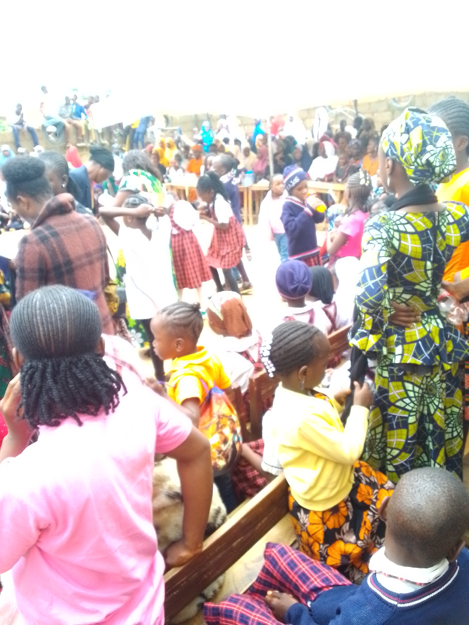 In an outdoor gathering in Unknown, Unknown, a diverse group of people, primarily children and women, are the focus. The scene is crowded with individuals engaged in various activities, though the specific nature of the event is not clear. Many are dressed in brightly colored clothing, with patterns ranging from plaid to floral. Several children are in the foreground, some wearing traditional African attire, while others are in more casual wear. One woman in the foreground is wearing a blue and yellow patterned dress. In the background, more people are visible, some sitting on benches, others standing. The environment appears to be a public space, possibly a market or a community gathering, with a wall and some vegetation visible in the distance. The lighting suggests it is daytime, with some overcast conditions.