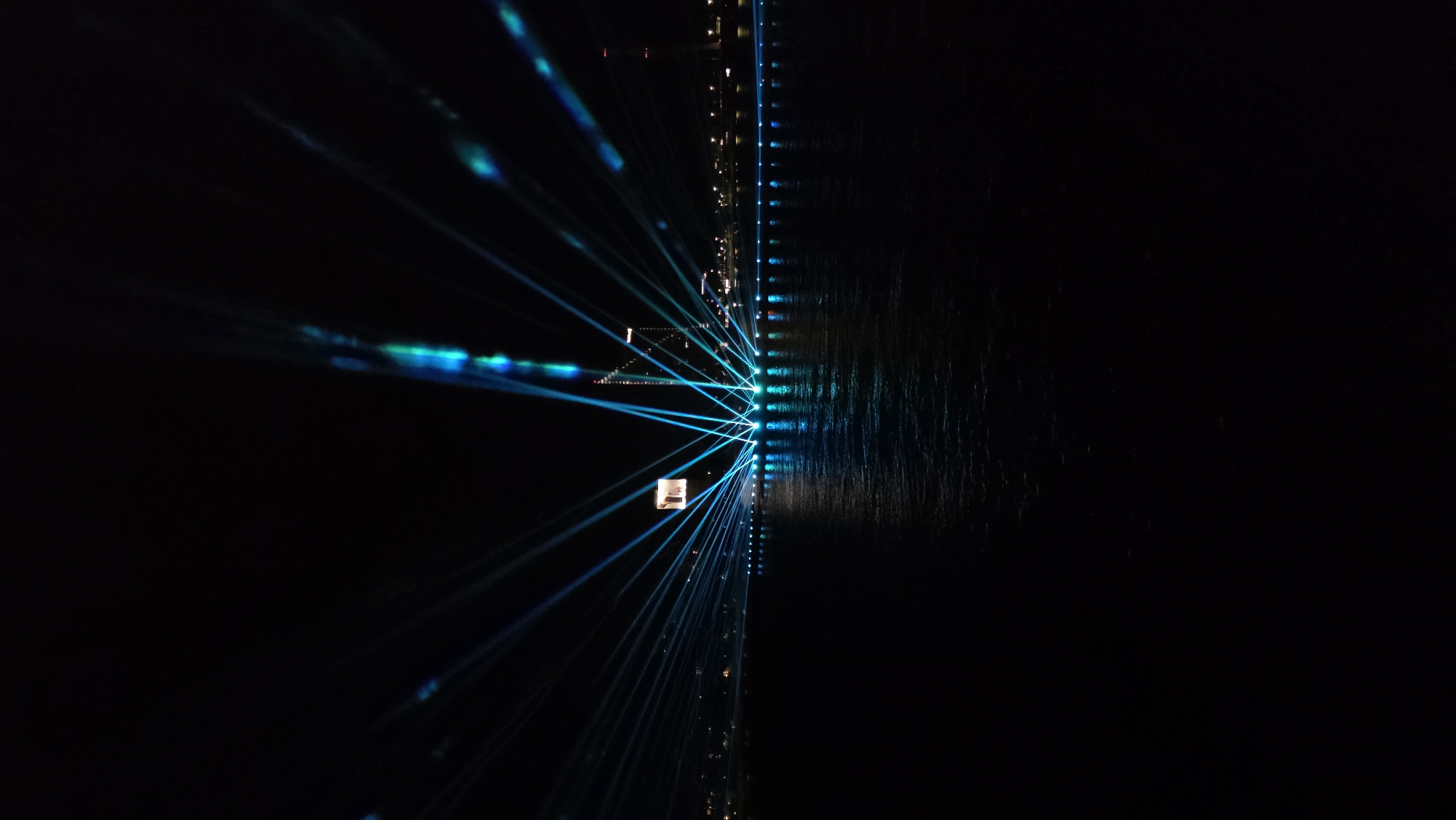 A nighttime cityscape features a body of water in the foreground, reflecting the light of a laser show and city lights. The water's surface is dark with ripples and highlights from the illumination. In the background, a line of lights suggests a city skyline, punctuated by a tall, modern building with a square illuminated sign on its side. A prominent laser show projects numerous blue and green beams from a low point on the waterfront, fanning out upwards and outwards into the dark sky. The lasers create a dramatic, radial effect, with some beams appearing to converge on the tall building. The overall impression is one of a vibrant, illuminated urban waterfront event. The scene depicts the Daugava River in Riga, Latvia, during an event.