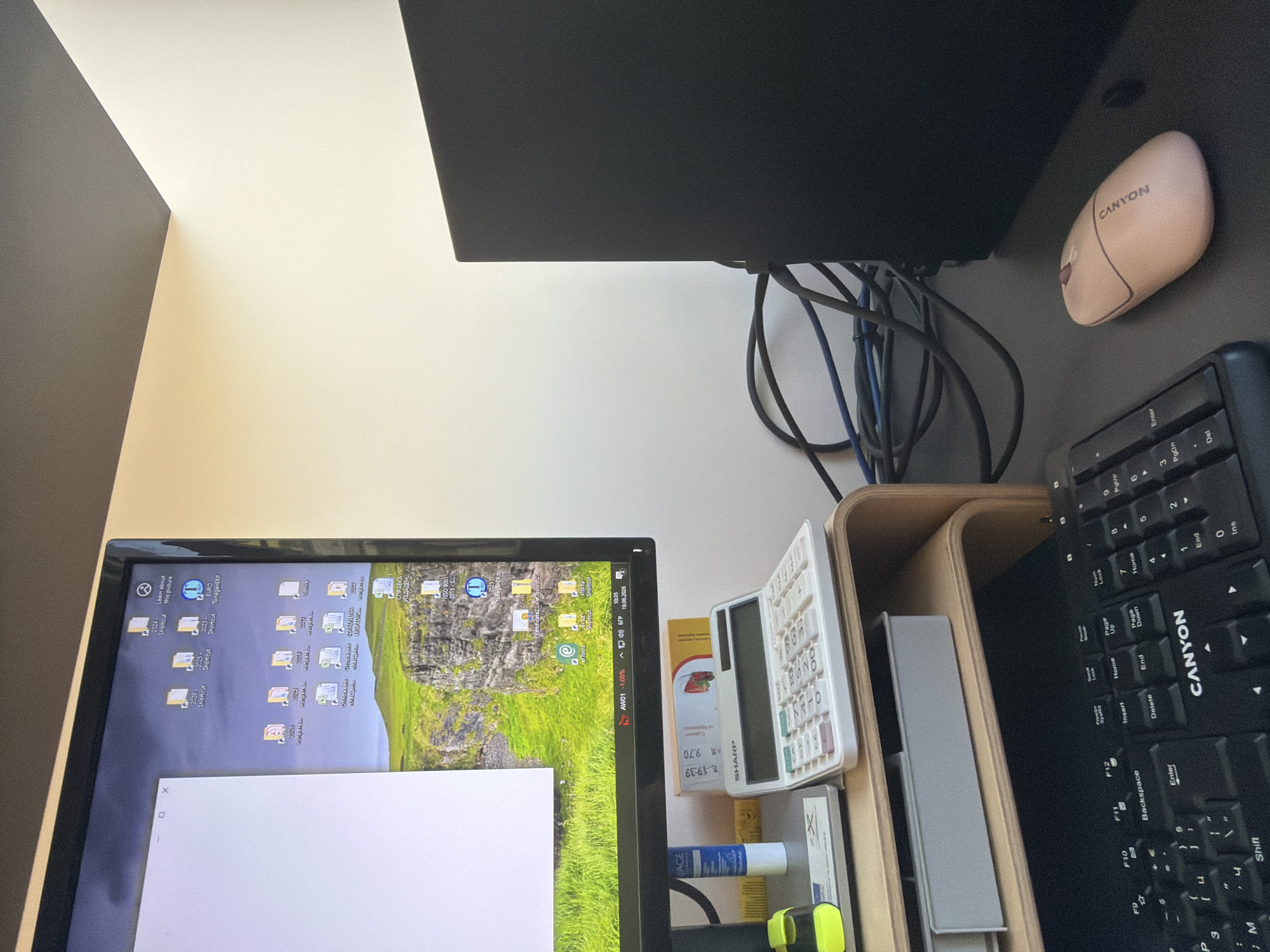 The image depicts a desk setup with a computer monitor displaying a desktop with various icons and a wallpaper of a scenic landscape. To the right of the monitor, there is a black computer tower and a tangle of cables. In the foreground, a black keyboard with the brand name "CANYON" is visible, and next to it, a pink computer mouse, also branded "CANYON". A white calculator is placed on a wooden monitor stand above the keyboard. A tube of glue and a yellow marker are also present on the desk. The background shows a plain wall with a dark ceiling. The scene is indoors, likely in an office or home workspace. The provided context states the location is Dolno Ablanovo, Bulgaria.