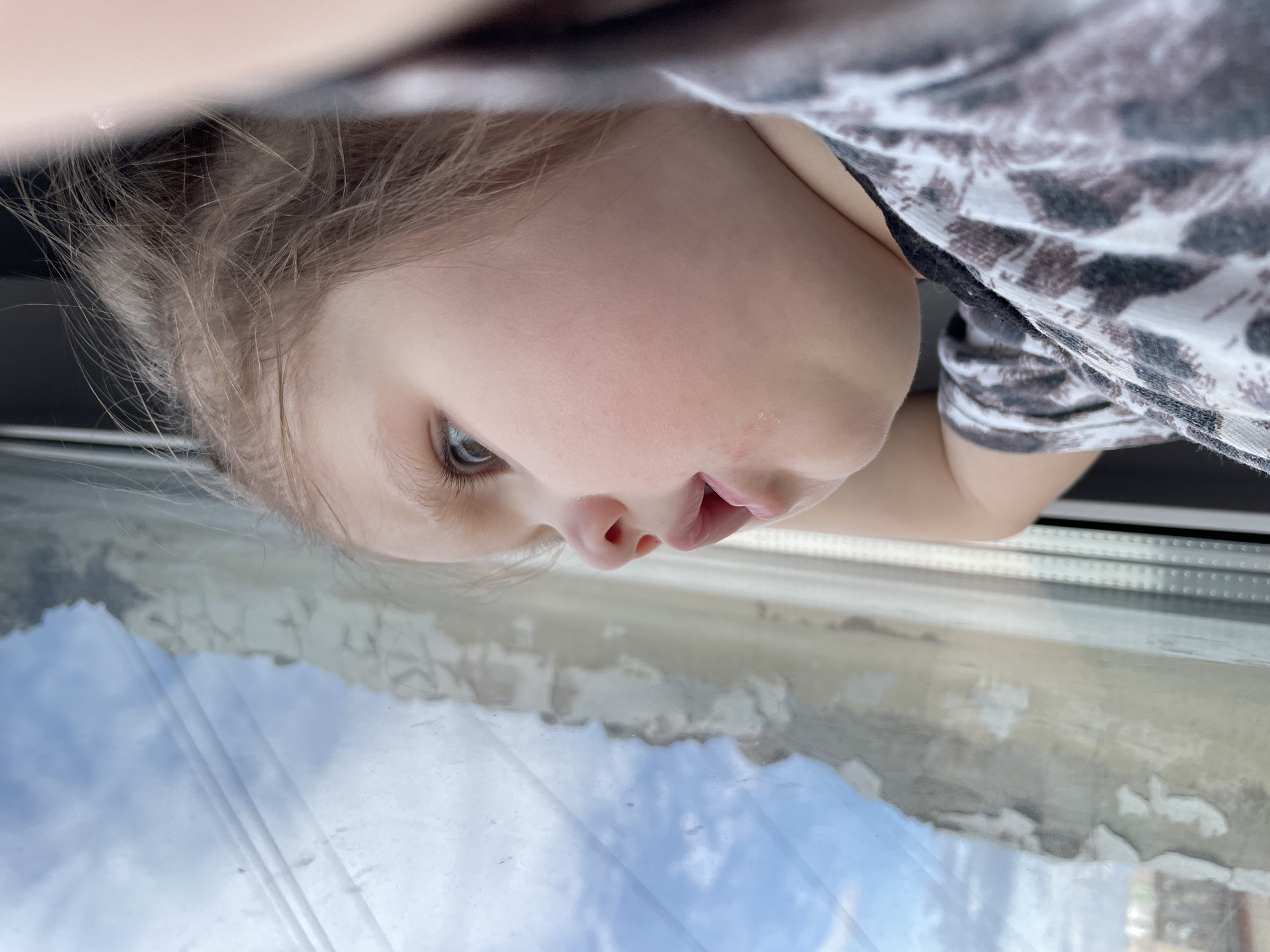 A close-up shot of a young child looking out of a window, with a view of the sky and clouds reflected on the glass. The child is wearing a patterned shirt. The peeling paint on the window frame suggests the location is a building in need of repair, potentially in the city of Izhevsk, Russia. The perspective is from above, looking down at the child.