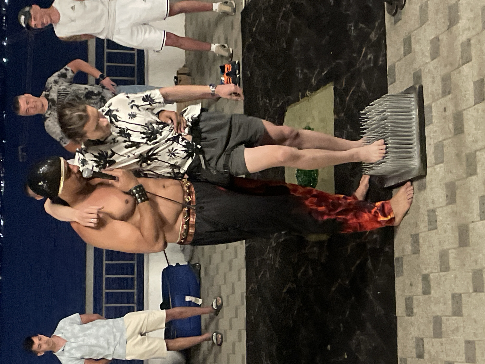 At an outdoor location with a body of water in the background, a man wearing a black head covering and a tribal-style belt is supporting another man's shoulder. The second man, dressed in a tropical-patterned shirt and grey shorts, is standing with one bare foot on a board covered in sharp nails. Several other men are visible in the background, some observing the scene and others conversing. The ground appears to be paved with square tiles.