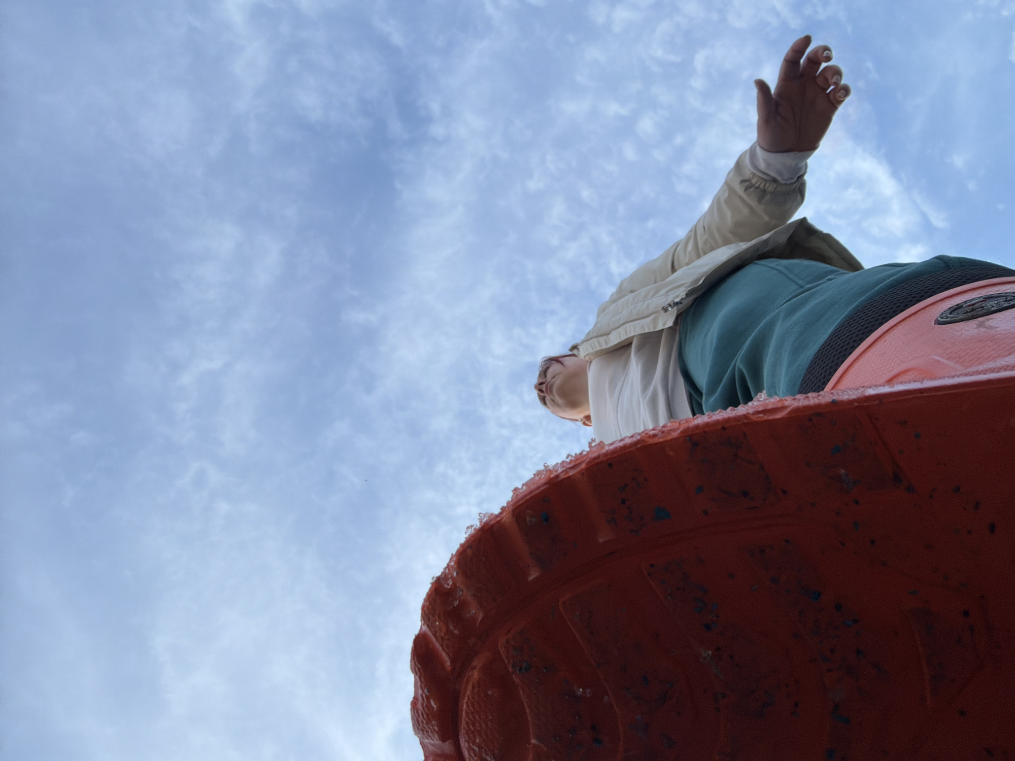 A low-angle photograph captures a person and a large orange-red object against a blue sky with scattered wispy clouds. The individual, positioned in the upper right, is visible from approximately the waist up. They wear a light-colored jacket over a lighter shirt and teal-colored trousers. Their right arm is extended upwards, with the hand open and fingers slightly spread, revealing light-colored fingernail polish. Their head is turned slightly to the left, showing short hair with subtle pink highlights. The lower foreground is dominated by a large, textured, orange-red object, consistent with the sole of a boot or specialized footwear, which features a black trim. Small crystalline particles, resembling ice or frost, adhere to the upper edge of this object, along with scattered dark debris. The exact location within the city of Chaikovsky, Russia, is not discernible from the visual information provided in the image.