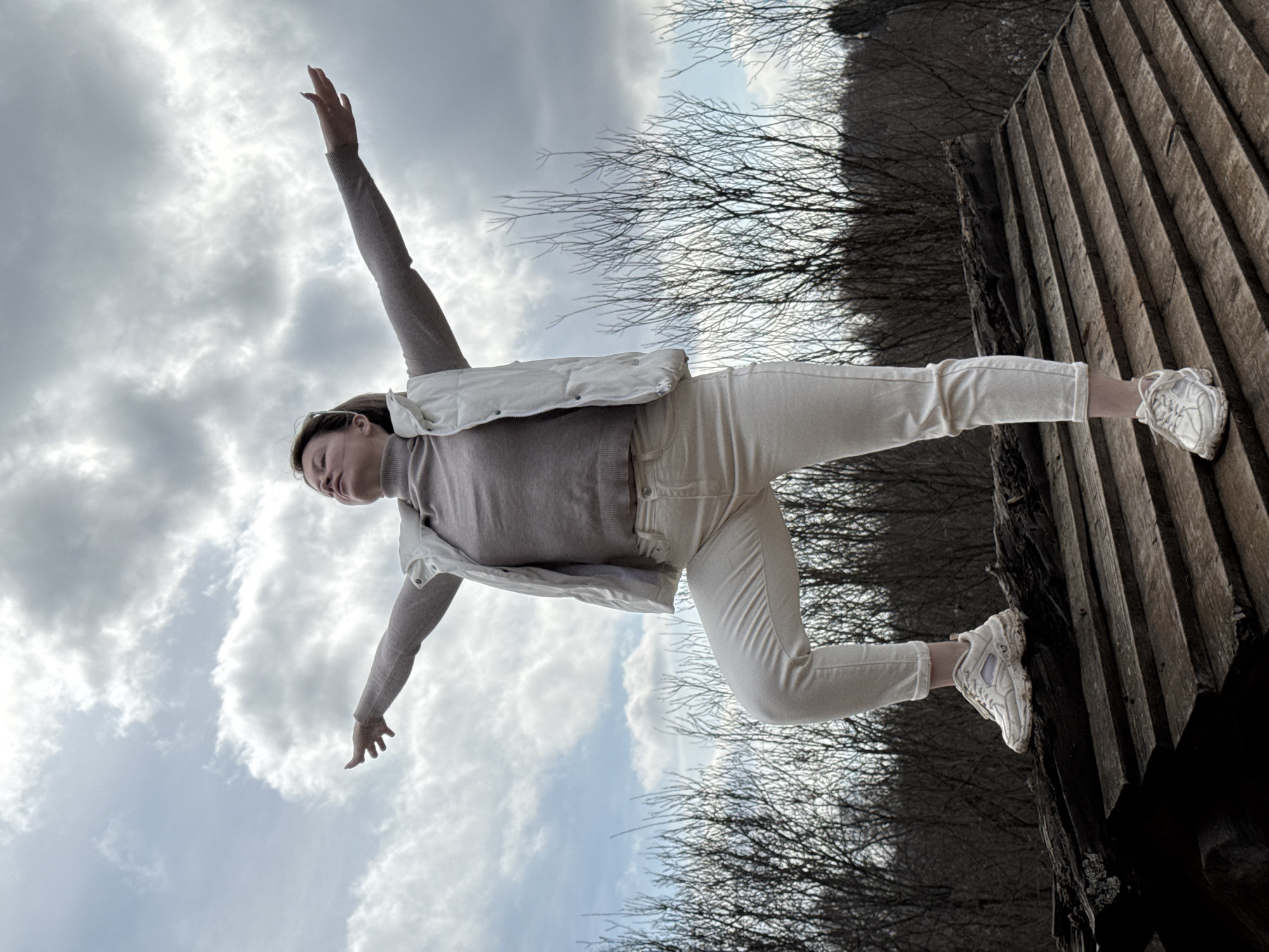 A young woman is depicted in a low-angle shot, likely taken in an outdoor setting. She is wearing a light-colored turtleneck sweater, white jeans, a white vest and white sneakers. Her left arm is extended upward towards the cloudy sky, while her right arm is extended downward. One of her feet is in contact with a wooden structure that resembles a bridge or walkway. The background features a forest of bare trees, also providing a hint of location. There is no readily accessible information to confirm Vassyata, Russia, as the location of this photo.