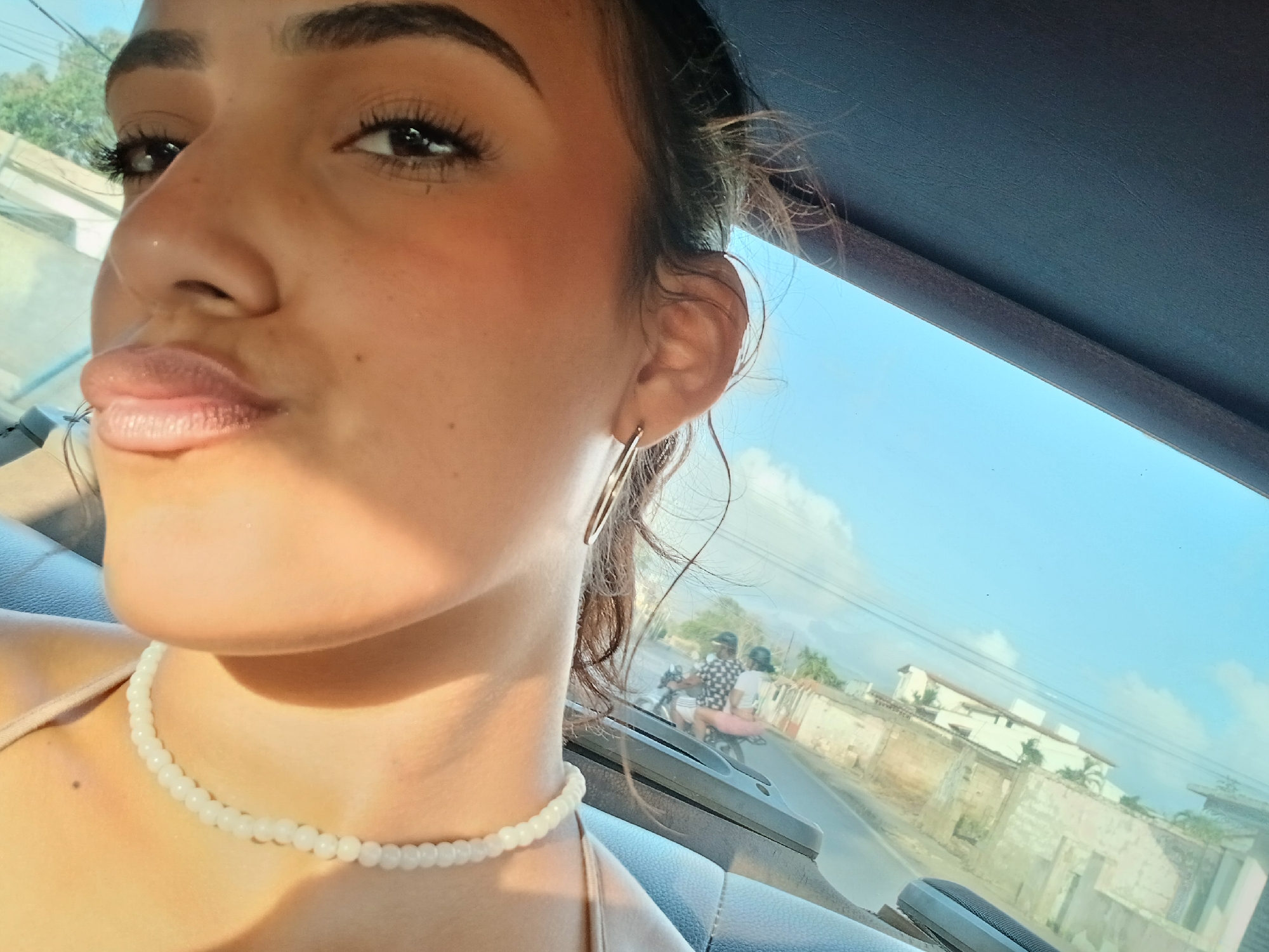 A close-up shot of a young woman's face, with her lips pursed for a kiss, fills the left side of the frame. Her dark hair is pulled back loosely, and she wears a delicate white beaded necklace and a thin hoop earring. Behind her, the blurred background reveals a street scene with two people riding a motorcycle, suggesting a location in a warm, possibly tropical climate. The bright sky and hints of buildings and trees in the distance further support this. The overall impression is a casual selfie taken from within a vehicle.