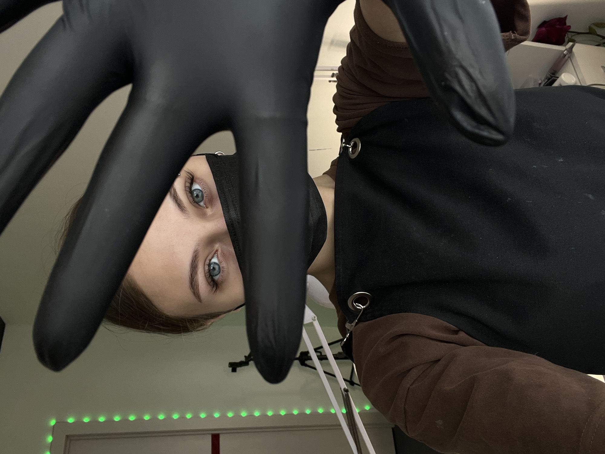 A young woman wearing a black face mask, brown long-sleeved shirt, and black apron is looking at the camera from a low angle, with a black-gloved hand obscuring part of her face. The room behind her features a strip of green LED lights and what appears to be a desk lamp. The scene does not provide enough information to confirm a location in the city of Фоки, Russia.