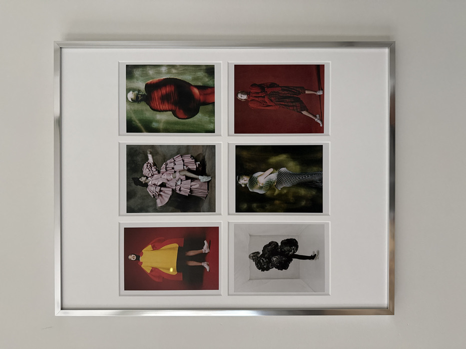 The scene is located in a building in the city of New York, United States of America.
A rectangular picture frame with a silver-colored metallic border is mounted on a light-colored, plain wall. Inside the frame, a wide white mat board frames six individual photographic prints, arranged in two rows of three.
**Top Row, Left:** A full-body portrait of a person wearing a voluminous yellow smock with oversized red sleeves, black shorts, and white sneakers, set against a solid red background. The person has dark hair and a black neck garment, with a discernible smile.
**Top Row, Middle:** A full-body portrait capturing a person in motion, wearing an elaborate pink gown featuring multiple ruffled tiers and large, sculptural sleeves. White sneakers are visible. The background is a dark, textured grey.
**Top Row, Right:** A full-body portrait of a person with light-colored hair, standing forward-facing. They wear a unique, form-fitting red garment with prominent horizontal pleats, creating a an abstract, elongated silhouette. The background is a mottled dark green and brown.
**Bottom Row, Left:** A full-body portrait of a person standing within a stark white, minimalist room with visible corners, suggesting a studio space. The person is adorned in a large, irregular black sculptural garment that significantly alters their body shape. Dark shoes are visible.
**Bottom Row, Middle:** A three-quarter portrait of a person viewed from the side, looking over their shoulder. They wear a black and white patterned, form-fitting dress with a structured, rounded element on the upper back/shoulders. The background is dark with subtle green and yellow tones.
**Bottom Row, Right:** A full-body portrait of a person striking a pose with one leg slightly extended. They wear a monochromatic red outfit consisting of a ruffled, high-necked blouse with puffy sleeves and wide, cropped plaid pants. White sneakers complete the ensemble. The background is solid red.