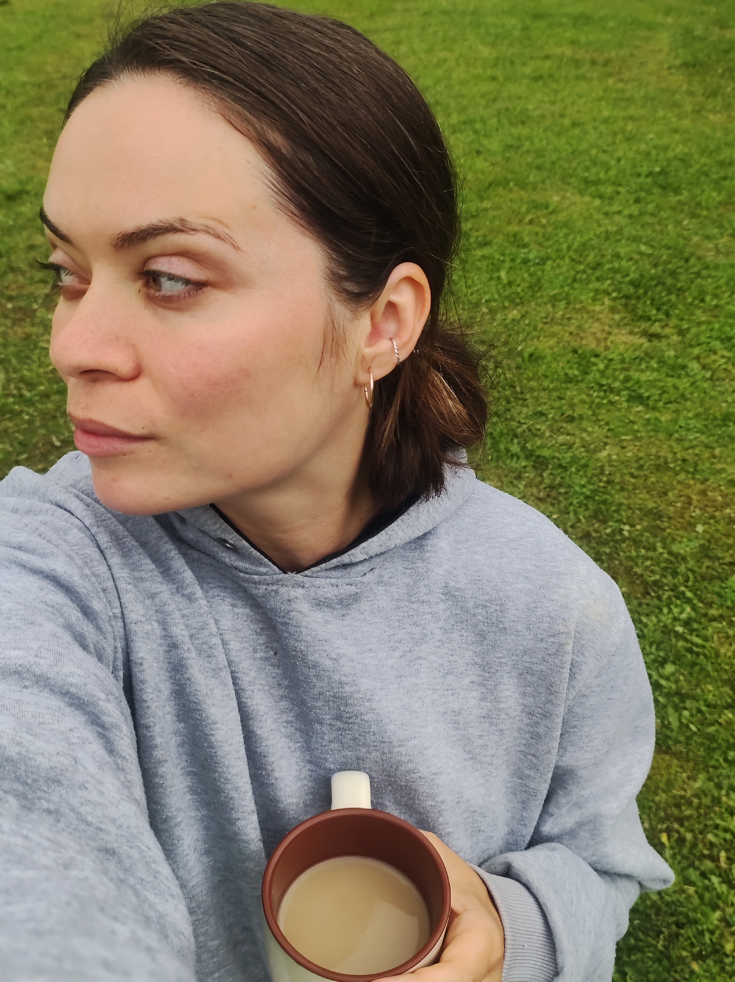 A close-up, chest-up selfie captures a female subject outdoors. She is positioned facing forward but looking sharply to her left, with her head slightly tilted down. She wears a plain light gray hooded sweatshirt. Her dark brown hair is pulled back, with wisps visible around her temples and ear. Her visible left ear features three piercings: a silver-toned twisted hoop in the helix and two small gold-toned hoops in the lobe. Her skin is fair with a slight natural flush on her cheeks. Her eyes are light in color. She holds a two-toned mug with a white handle and a brown rim, containing a light brown, milky beverage. The background consists of out-of-focus green grass, indicating an outdoor environment. The image provides no visual information to identify a specific city or country, such as Фоки, Russia.