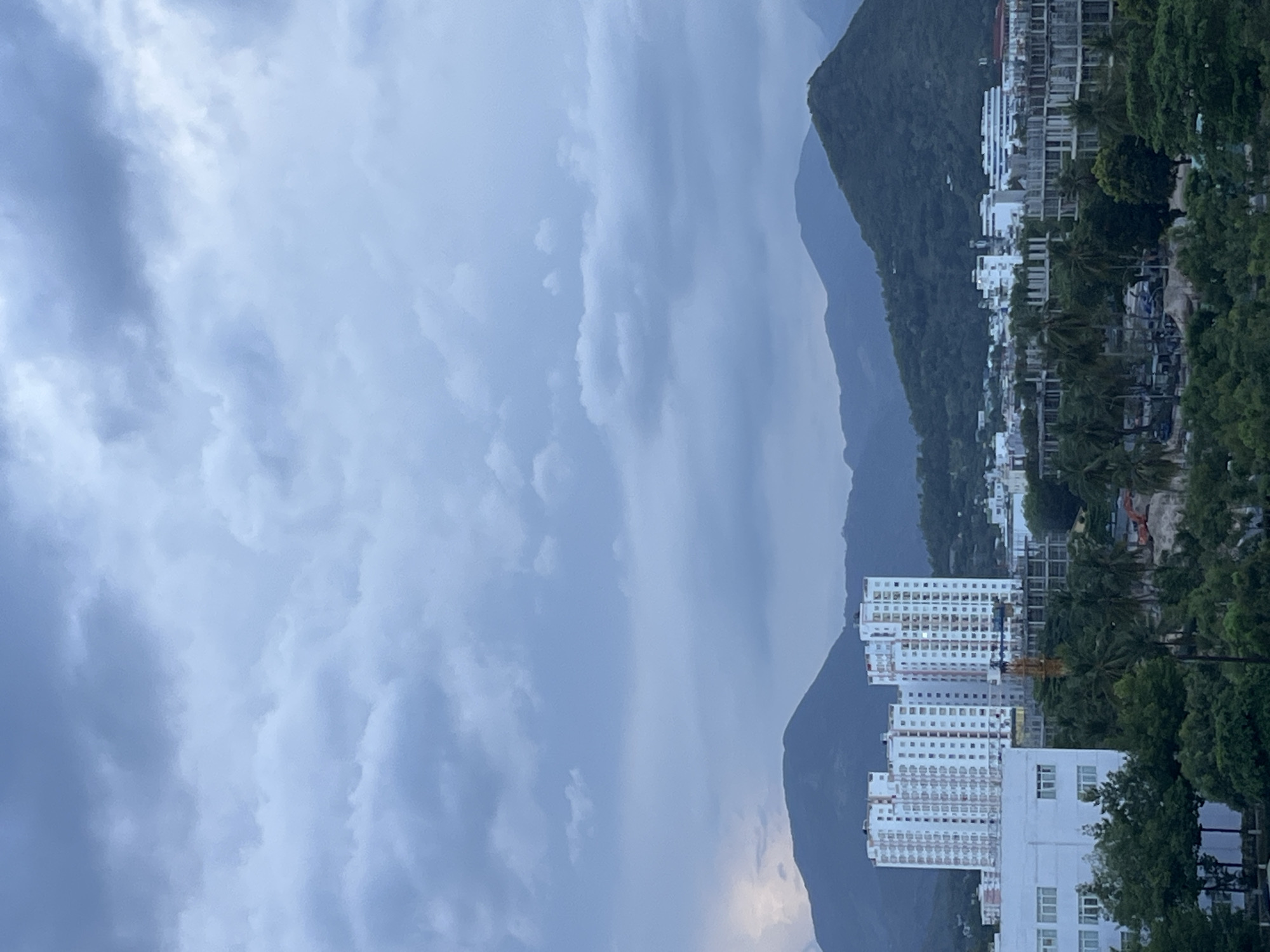 The image shows a cloudy day in Bac Nha Trang Ward, Vietnam. In the foreground, tall, white apartment buildings rise towards the sky. Behind these buildings, a green, mountainous landscape provides a backdrop. The sky is filled with heavy, grey clouds, giving the scene a muted, overcast appearance. The city's built environment contrasts with the natural surroundings of the mountains. The photograph seems to be taken from an elevated position, offering a panoramic view of the area. There is no evidence of human activity in the image.