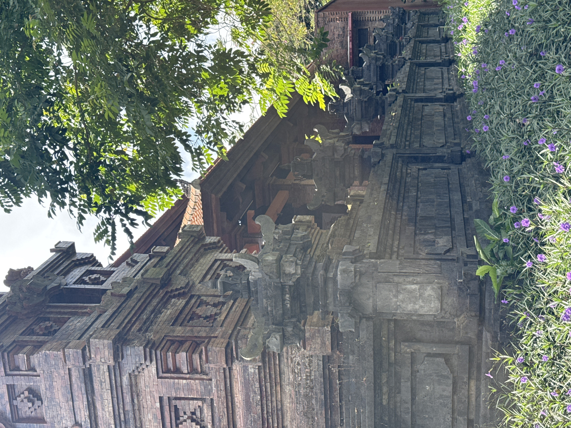 The image shows a side view of a building with intricate brickwork and carved stone accents. The architecture appears to be traditional, possibly a temple or a historically significant structure. In the foreground, there is a lush bed of green plants dotted with small purple flowers. To the right and above the building, dense green foliage from trees overhangs the scene, with dappled sunlight filtering through the leaves. The building's facade features layered brickwork with decorative patterns and detailed stone sculptures that adorn the wall and corner structures. The overall impression is of a serene outdoor setting with a focus on the architectural details and surrounding nature. This building is located in Kuta Selatan, Indonesia, a region known for its cultural heritage and natural beauty.