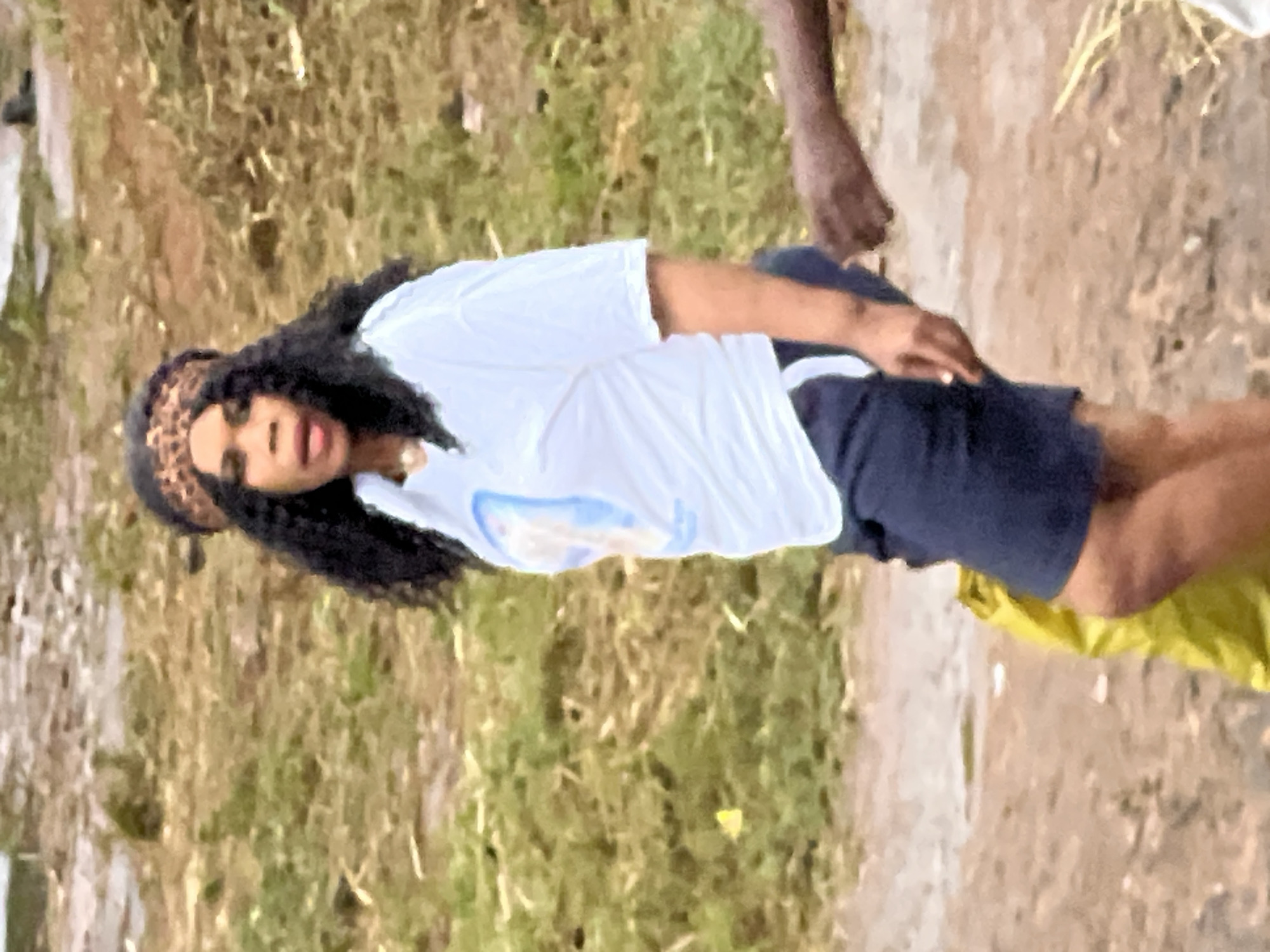 In Nkpor, Nigeria, a woman with dark, curly hair and a leopard-print headband stands on a dirt path. She is wearing a white t-shirt with a blue and white graphic on the front and dark blue shorts with a white stripe. A yellow bag rests on the ground next to her left leg, and a person's arm is visible to her right. The background consists of dry, yellowish grass and scattered debris, suggesting an outdoor, unpaved area.