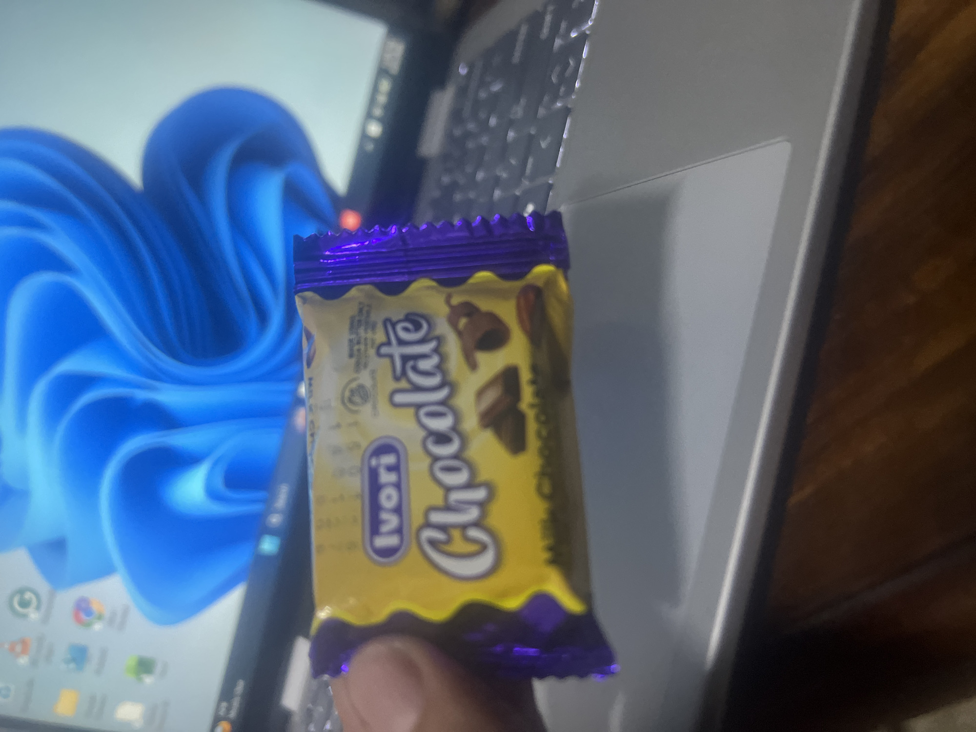 A close-up shot features an Ivori Milk Chocolate candy bar, identifiable by its yellow and purple packaging and the "Ivori Chocolate" branding. The candy bar is held in someone's hand, with the person's fingers visible. The background consists of a laptop, partially showing the keyboard, trackpad, and the Windows operating system display. The laptop is resting on a wooden surface. The location in Dar es-Salaam, Tanzania is not discernible from the visual information provided.