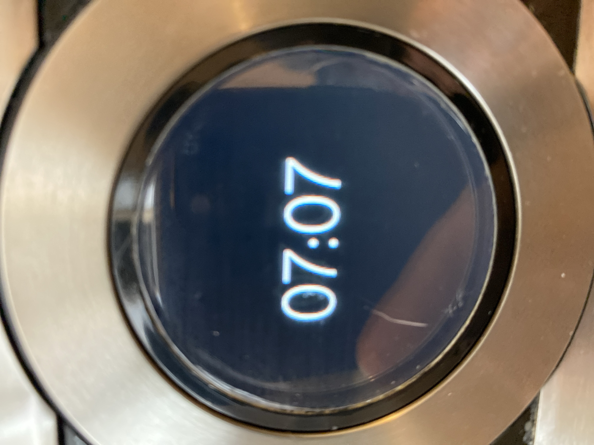 A close-up photograph shows a round, digital display screen surrounded by a metallic bezel. The screen displays the time "07:07" in a vertically oriented format. The background of the screen is dark, and the numbers are illuminated in a light blue or white color. There is a scratch or crack visible on the screen. The location is not discernible from the visual information provided.