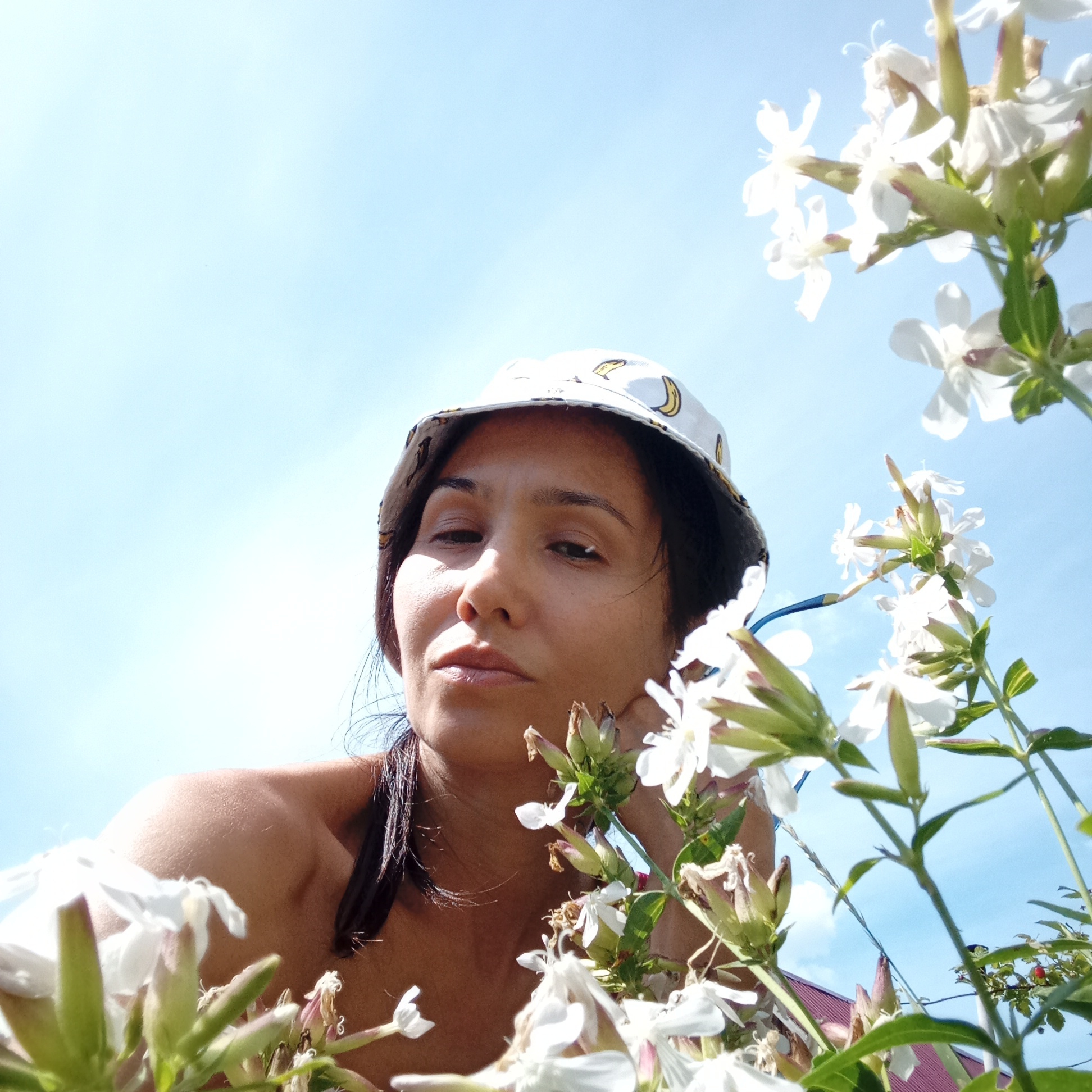 A low-angle, waist-up, medium shot shows a woman looking towards the camera. She is wearing a white bucket hat with a banana print. The woman has dark hair, which is partially visible under the hat and falling onto her shoulders. Her shoulders and upper chest are exposed, suggesting she is not wearing a shirt. The background is a bright blue sky with white flowers in the foreground and to the right of the woman. The woman's face is illuminated from above, casting some shadows on her features. She appears to be outdoors.
