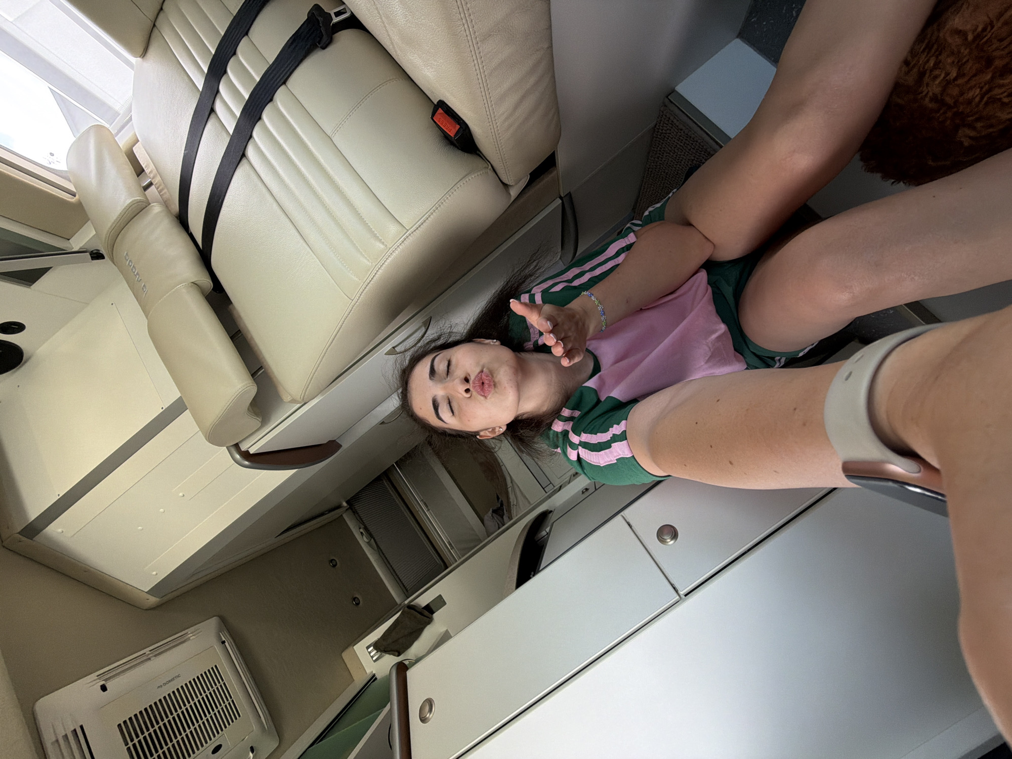 In the image, a young woman is sitting inside a vehicle, likely a campervan or RV. She is wearing a green t-shirt with pink stripes and green shorts. She is puckering her lips and holding out her hand as if blowing a kiss. Her eyes are closed. The interior of the vehicle is visible, featuring cream-colored leather seats, white cabinetry, and an air conditioning unit on the ceiling. The scene appears to be a selfie taken by the woman. There is no indication of the location being the city of Nea Moudania, Greece.