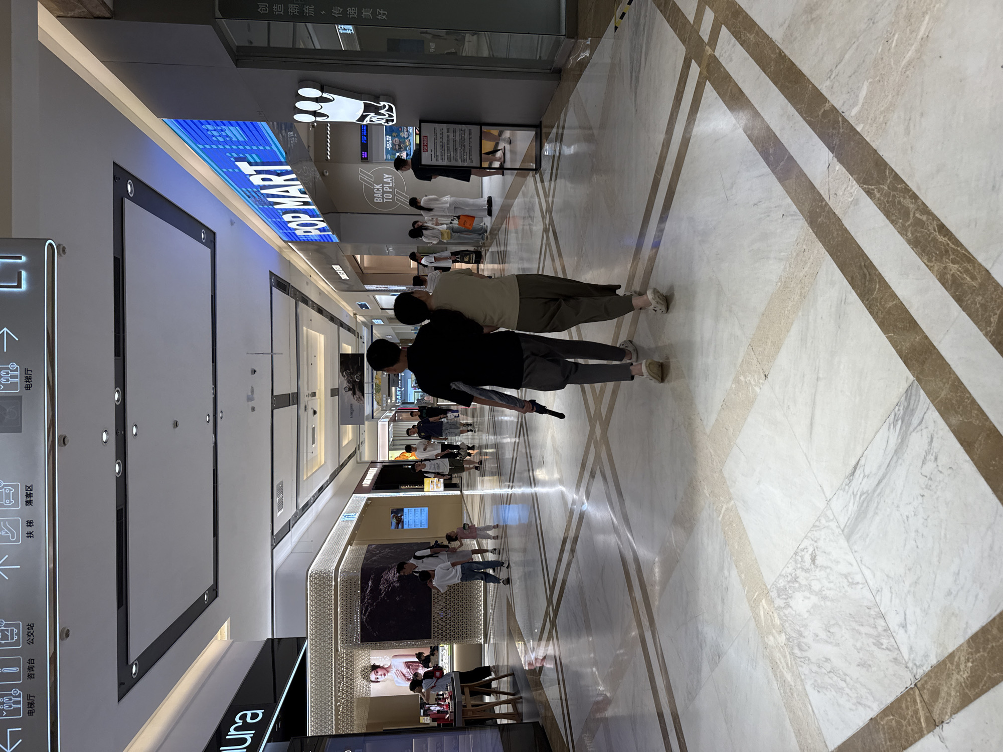 This image depicts a brightly lit indoor shopping mall, likely in Huizhou, China, with a focus on a storefront for "POP MART" and the people walking through the hallway. The setting is a modern mall with polished marble floors featuring geometric patterns and high ceilings. The ambient light suggests it could be daytime, as evidenced by the natural light filtering in from above and the overall illumination.
The main subjects are two individuals in the foreground, seen from behind and walking away from the camera, their arms linked. They are casually dressed, suggesting a relaxed atmosphere. Further down the mall, other shoppers are visible, some browsing, some walking, and some queuing near the POP MART store. The store itself is a prominent feature, with a large, brightly lit sign displaying "POPMART" and the tagline "BACK TO PLAY." A display sign nearby lists store information, and another large screen shows what appears to be promotional content. The presence of directional signs for "电梯厅" (Elevator Hall) and "公交站" (Bus Stop) indicates the mall's accessibility and services.
The overall mood is one of everyday commerce and leisure, with people going about their shopping or simply enjoying a walk through the mall. The clean, well-maintained environment and the presence of popular retail brands suggest a typical urban commercial space. There are no overt indications of a specific event or time of day beyond general mall activity.