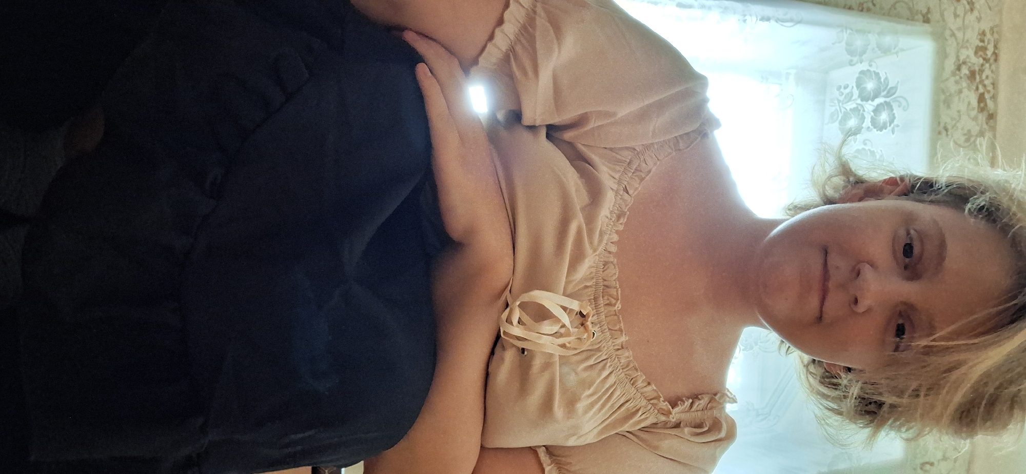 The image shows a medium shot of a person from the chest up, sitting indoors. The person has short, light-colored hair and is looking directly at the camera with a neutral expression. They are wearing a light beige, off-the-shoulder top with ruffled details and a lace-up front. Their left arm is resting on their lap, which is covered by a dark blue skirt. The background consists of a window with a sheer curtain that has a floral pattern. The lighting suggests it is daytime, with natural light coming through the window. The scene is devoid of any specific identifiable landmarks that would place it within Petropavl, Kazakhstan.