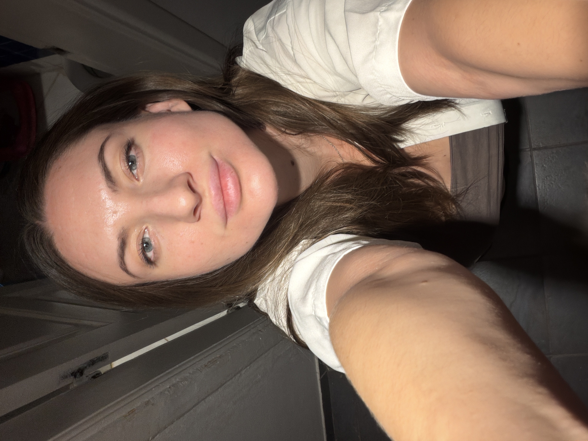 A young woman with long brown hair and blue eyes is taking a selfie. She is wearing a white collared shirt over a brown tank top. Her skin appears flushed, and there is a slight sheen from what could be sweat or moisturizer. She is indoors, in what appears to be a dimly lit hallway or bathroom with tiled flooring and textured walls. The lighting suggests artificial light is being used, casting shadows and highlighting her features. There are no other people or significant objects visible in the frame, besides the implied background elements of the indoor space. The image does not provide any specific clues or context that would place the scene in Sharm El Sheikh, Egypt; it depicts a generic indoor setting.