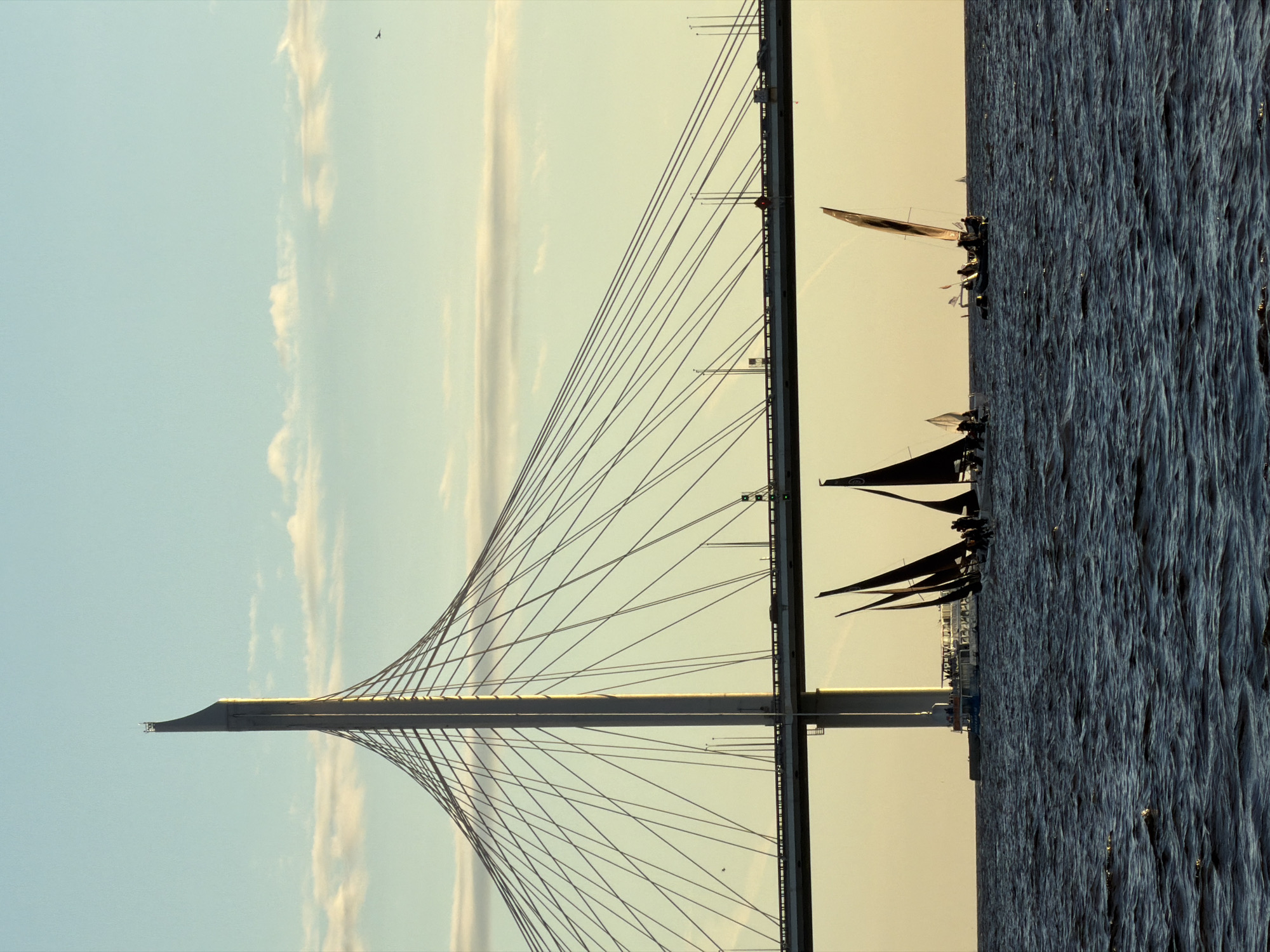 The image captures a scene on the Gulf of Finland, with the Western High-Speed Diameter bridge, a prominent landmark in Saint Petersburg, Russia, dominating the upper portion of the frame. The sky is a clear blue with scattered, wispy clouds tinged with the warm hues of sunset. Below the imposing structure of the cable-stayed bridge, several sailboats with dark sails are navigating the choppy, dark blue water. A larger white passenger boat is visible in the background near the bridge's support. The overall atmosphere suggests a late afternoon or early evening in a coastal urban environment.