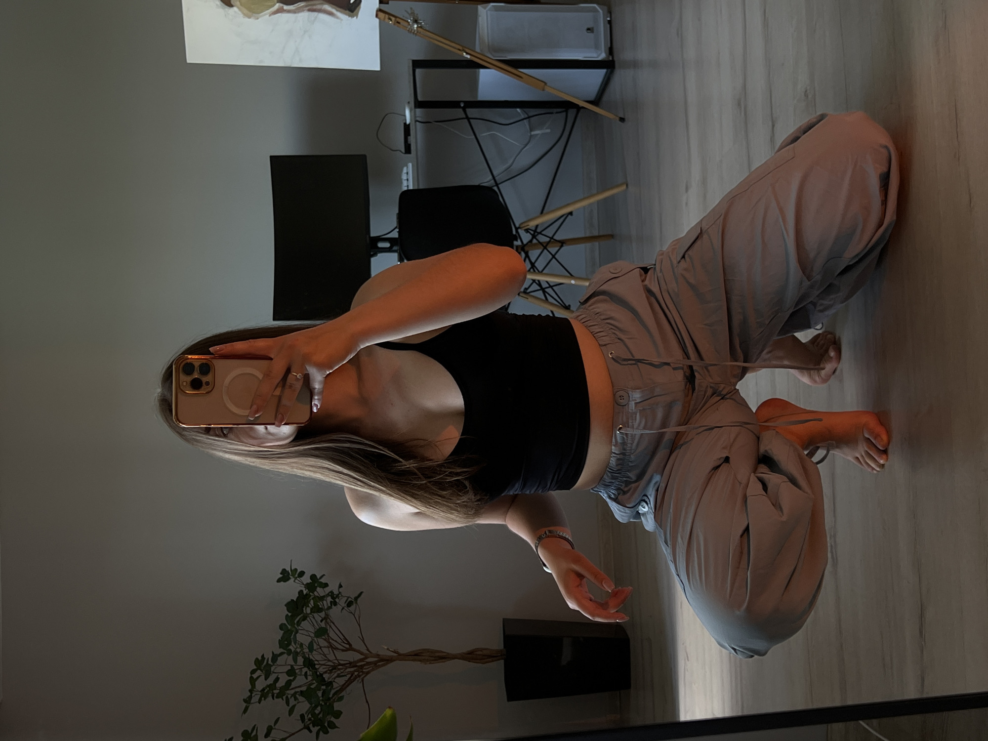 A young woman is taking a mirror selfie in a room, likely her home, in Ternopil, Ukraine. She is wearing a black tank top and light gray cargo pants. She is crouching down and holding her phone, which has a gold case, up to the mirror. In the background, there is a plant in a black pot, a desk with a computer monitor, a chair, and an easel with a painting. The room appears to be a living area or bedroom. The lighting is dim with warm tones, possibly from artificial light.