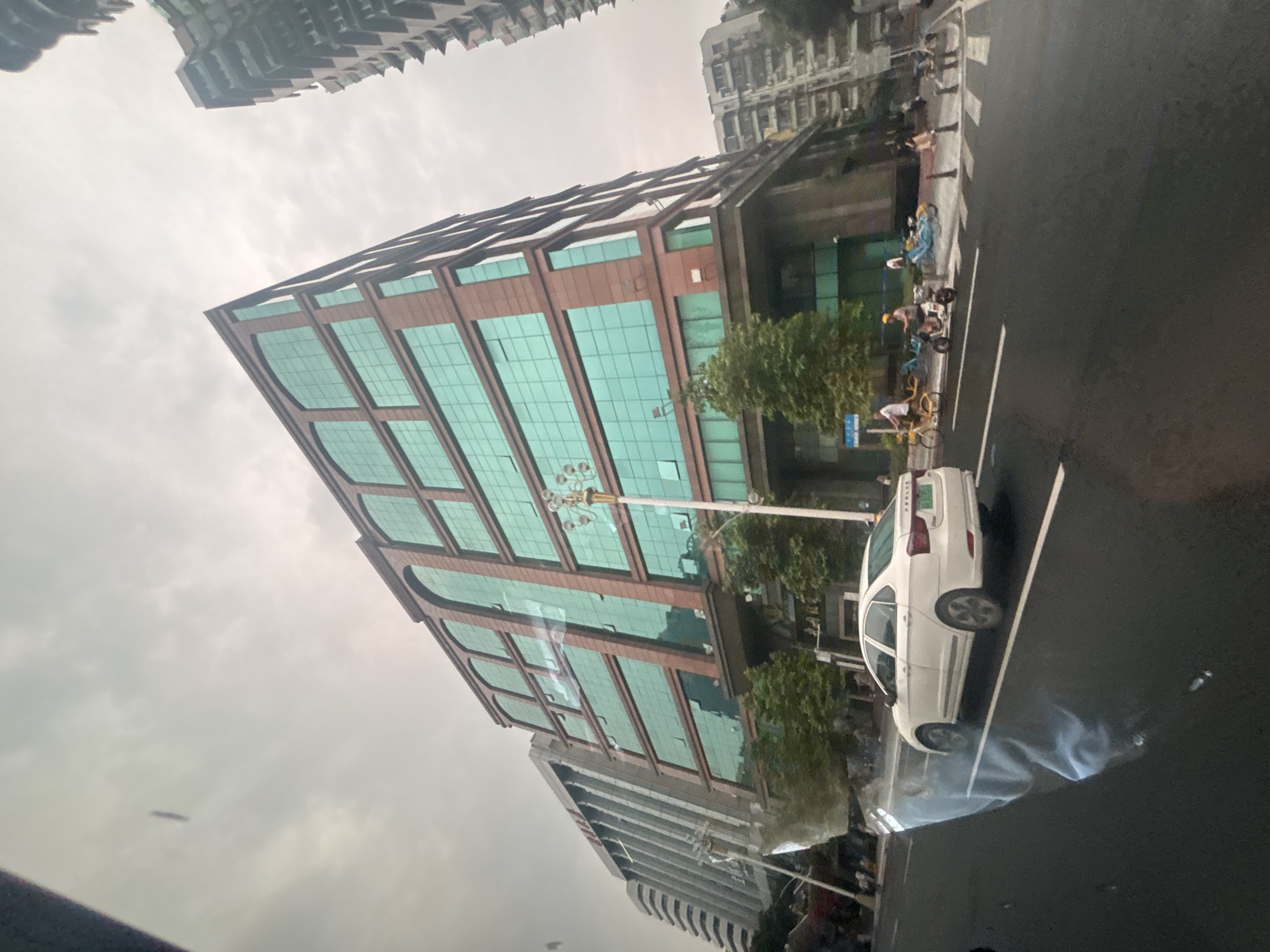 This is a street-level, slightly upward-angled photograph taken from a vehicle's perspective, looking towards a modern, multi-story building in Yuexiu District, China. The building has a distinctive red-brown facade with large, repeating arched windows filled with teal-tinted glass. Multiple stories of these windows are visible, suggesting a commercial or office building. To the left of this main building, a taller, lighter-colored building with a more utilitarian design is partially visible. The sky is overcast, with a hazy, diffused light.
In the foreground, a white sedan is driving on the asphalt road. To the right of the road, a sidewalk area features several people and electric scooters. One person is riding a yellow bicycle, and another is on a white electric scooter. Other individuals are standing on the sidewalk, and several blue and yellow shared electric bikes are parked. A lamppost with multiple lights stands near the main building. The overall scene depicts a typical urban street environment with contemporary architecture and common modes of transportation.