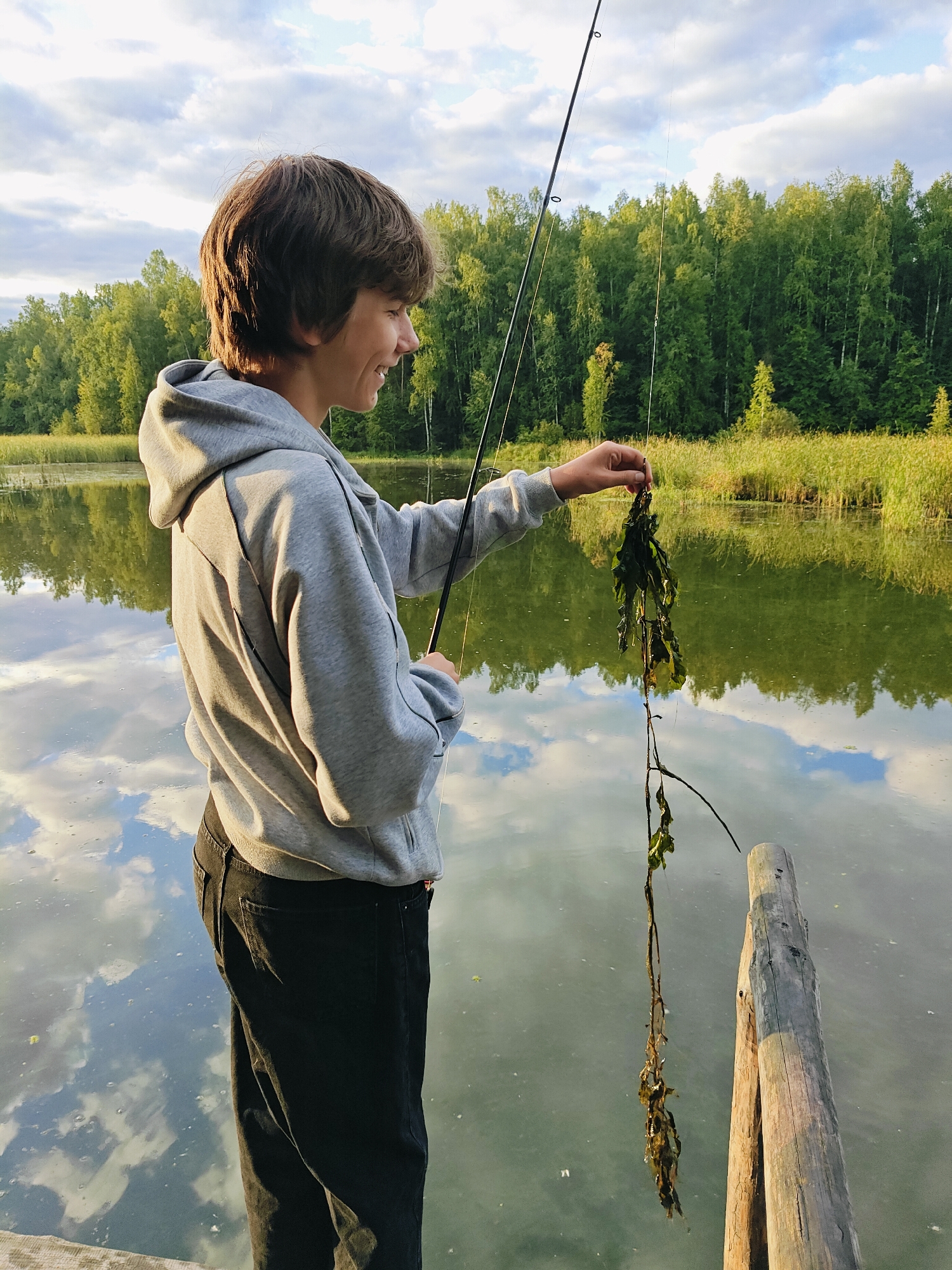 In Chaikovsky, Russia, on a calm body of water, a young person stands on a wooden pier, holding a fishing rod. They are smiling and looking at a tangle of dark green aquatic plants they have just reeled in. The water reflects the cloudy sky and a dense forest that lines the opposite bank. The sunlight casts a warm glow on the scene, highlighting the texture of the wooden pier and the youthful face of the angler.