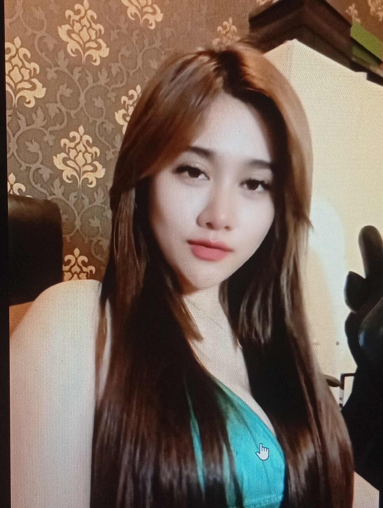 A young woman with long, light brown hair, parted in the middle, is positioned centrally in the frame. She is looking directly at the camera with a neutral expression. She is wearing a teal-colored garment. The background consists of a patterned wallpaper in shades of brown and gold, with floral motifs. A black object, possibly a chair or clothing, is partially visible to her right. The image appears to be taken indoors. There is a cursor icon overlayed on the woman's chest. This image is not specific to Lhokseumawe, Indonesia, as no identifying landmarks or contextual clues are present.