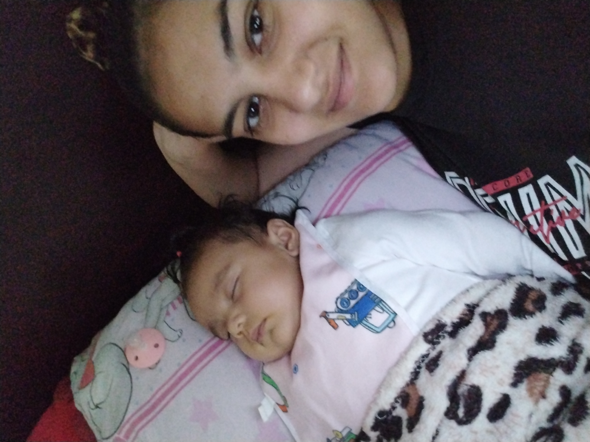 The image shows a person and a baby lying on what appears to be a bed or sofa. The person, likely an adult female, is positioned at the top of the frame, smiling at the camera. She has dark hair pulled back and is wearing a dark shirt with white lettering. Below her is a baby, asleep on a pillow with a pink pacifier attached. The baby is partially covered with a pink blanket with an illustration of a blue vehicle and a furry animal-print blanket. The background is a dark brown color. The location is unknown.