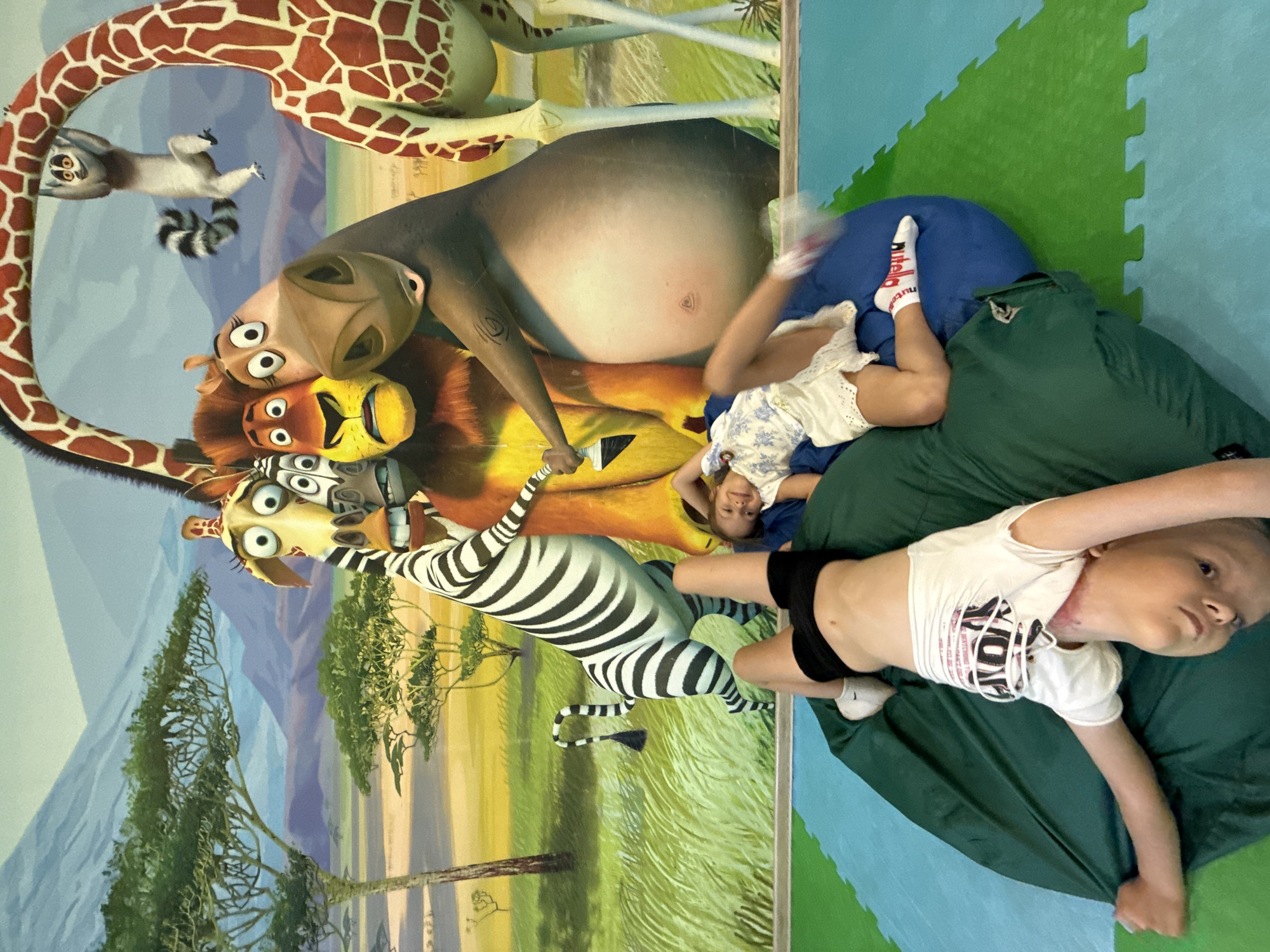 In an indoor recreational area, two children lie on large beanbag chairs in front of a mural depicting characters from the animated movie "Madagascar." A boy in black shorts and a white t-shirt is on his back on a green beanbag, with his head and upper body hanging upside down. A girl in a white floral top and shorts is on her back on a blue beanbag next to him, with her legs bent and one foot up. The mural shows the main characters of the movie, including a zebra, a lion, a giraffe, and a lemur. The floor is covered with interlocking mats in blue and green.