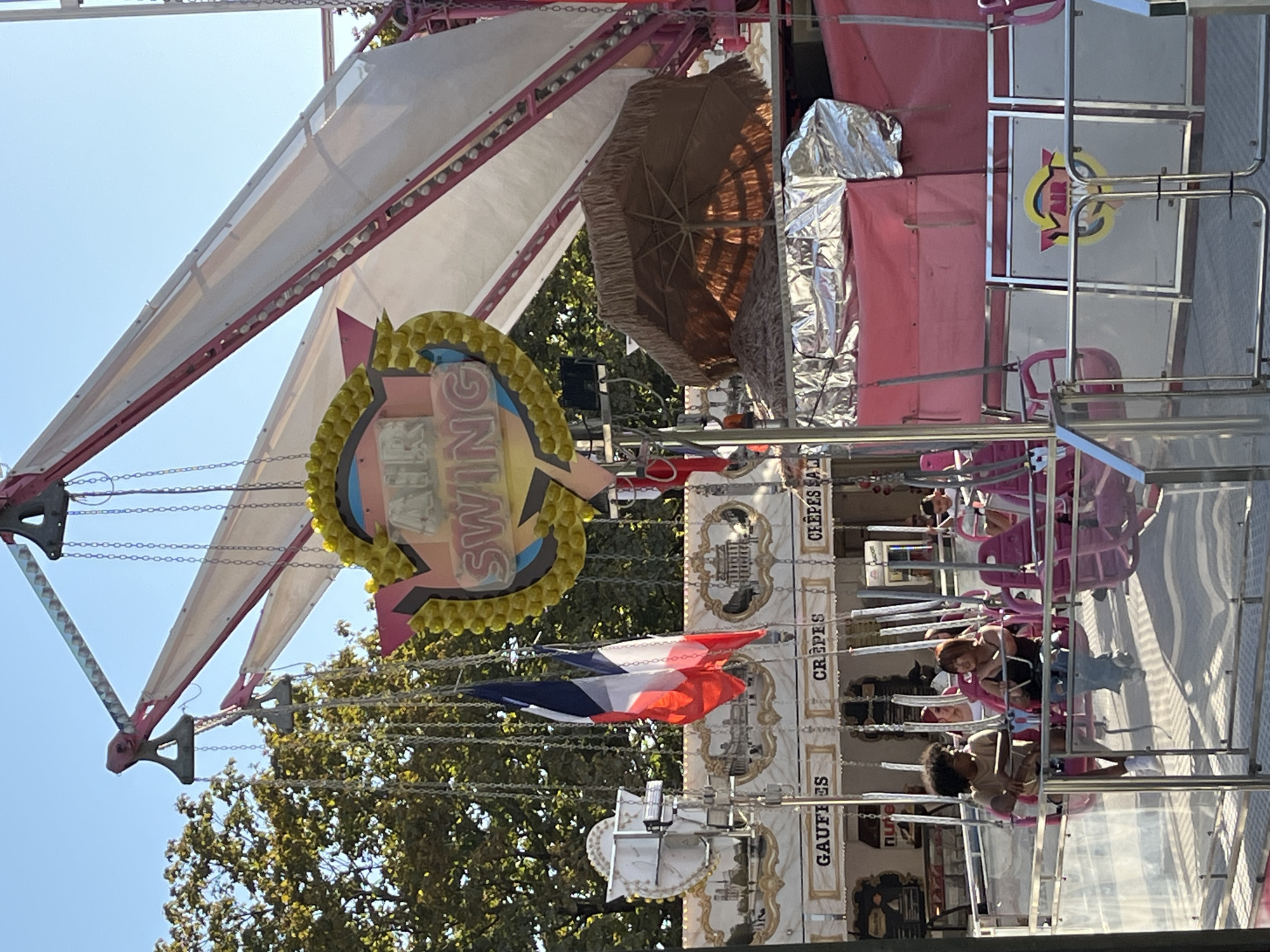 The image depicts a scene at a fairground or amusement park in Paris, France, likely during the daytime given the clear blue sky. The central focus is a carnival ride labeled "AIR SWING" with a distinctive yellow, circular emblem. The ride features multiple chains suspended from a tall, pink and white structure, with individual seats attached to the chains. Two French flags, one larger than the other, hang from chains on the left side of the ride.
In the foreground, several people are seated on the ride's pink seats, some appearing to be in mid-swing. Two young individuals, one with curly dark hair and wearing a tan t-shirt, and another with short dark hair and a black top, are prominent in the lower left section of the frame. Other fairgoers can be seen further back on the ride.
Below the ride, a concession stand area is visible, with signs indicating "GAUFFES" (waffles), "CREPES," and "CREPES SALEES" (savory crepes). The architecture of the concession stands suggests a European carnival aesthetic, with ornate framing and detailed murals depicting buildings. A brown, fringed umbrella is visible on the right side of the image, providing shade. The ground appears to be a paved surface, typical of a fairground. The overall atmosphere suggests a lively outdoor event.