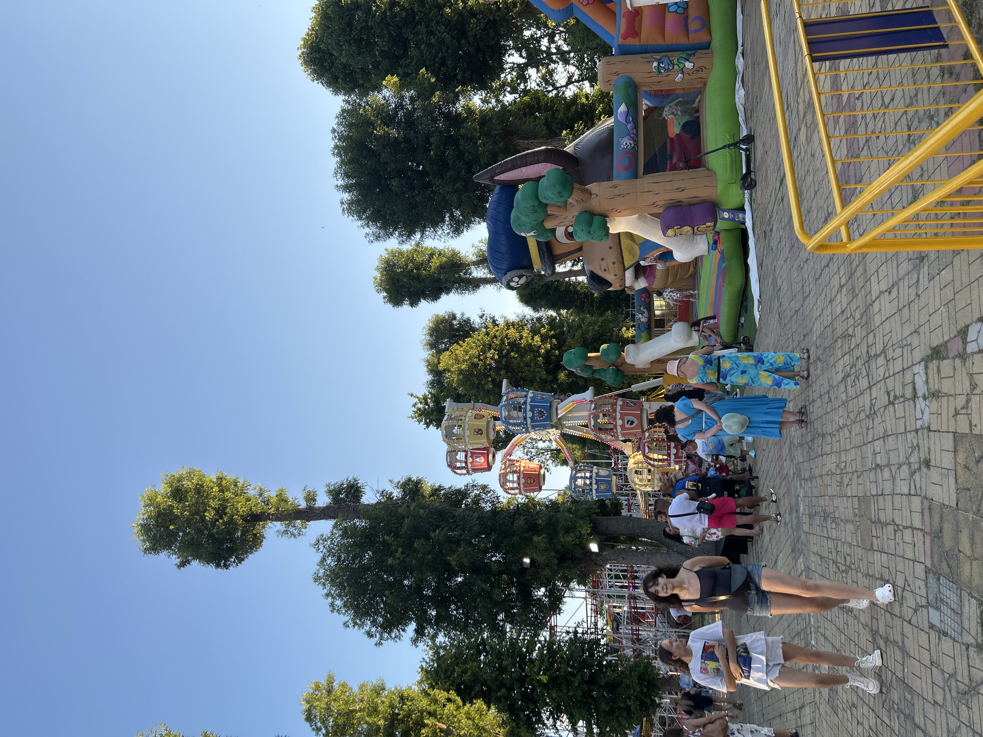 In Unknown, Unknown, a sunny outdoor amusement area features a Ferris wheel with colorful gondolas and a large inflatable obstacle course designed to look like a dog character. Several people are walking on a brick-paved walkway in the foreground. Two young women are walking towards the camera, one in a white t-shirt and shorts, the other in a black tank top and denim shorts. Behind them, other park-goers are visible, including a man in a white t-shirt and pink shorts, and a woman in a blue dress. The background is filled with tall green trees and a clear blue sky.